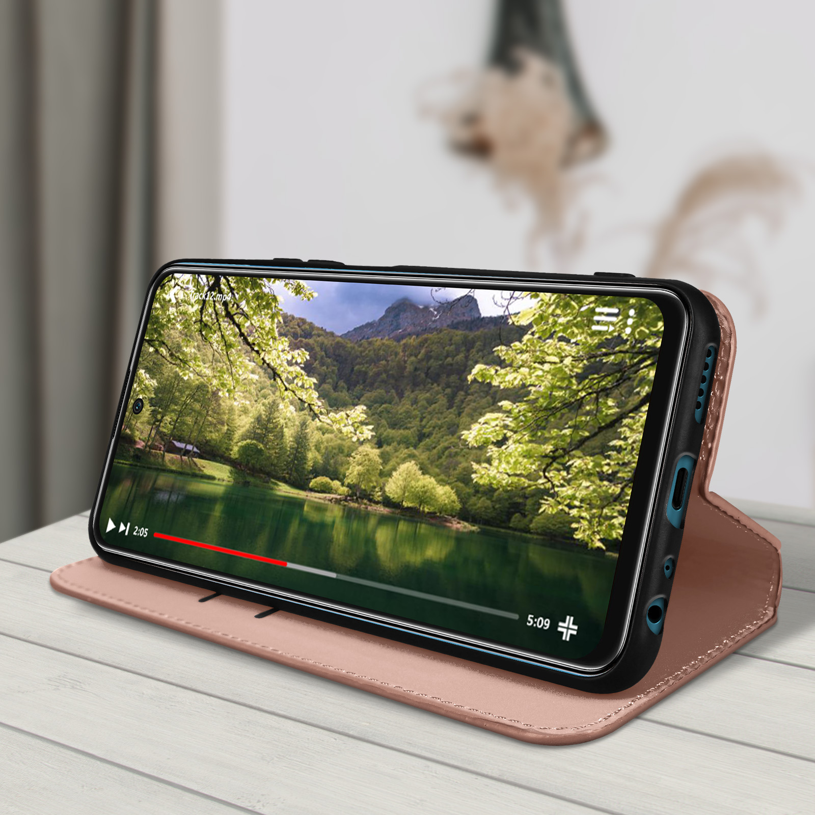 Un telefono in una custodia rosa è aperto, mostrando un video. Lo sfondo ha un tavolo di legno e un muro sfocato.