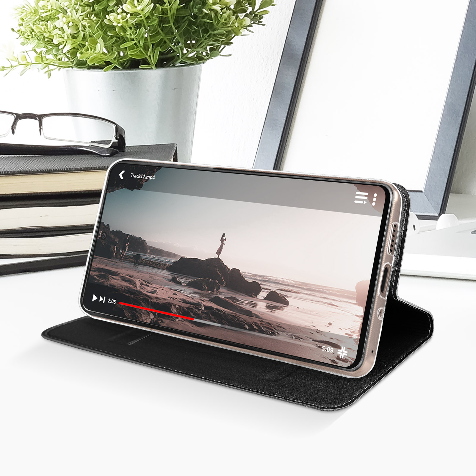 Un telefono mostra un video su una custodia nera, con una scena di spiaggia, su una superficie bianca.