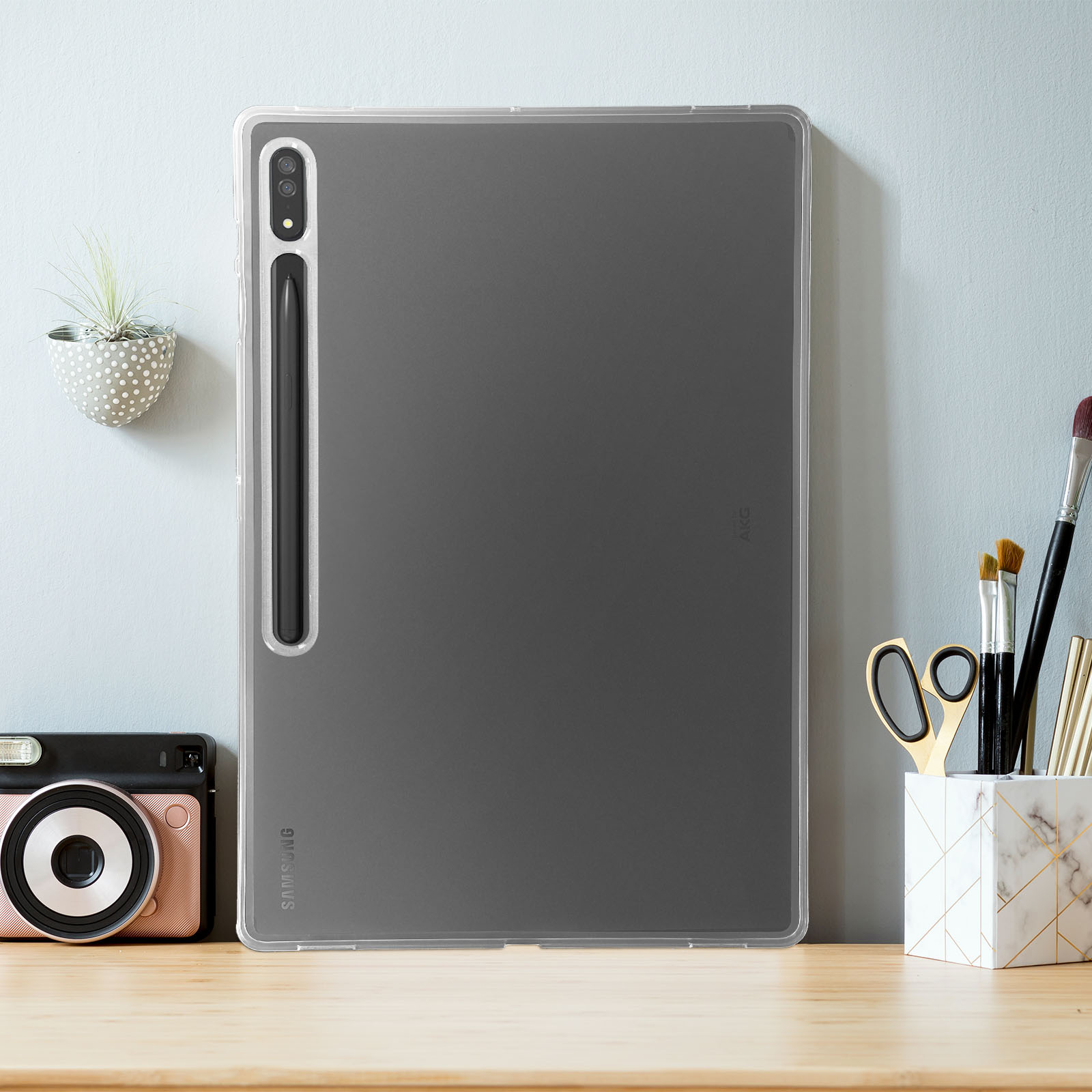 Tablet in klarer Hülle auf Holztisch, mit Schreibtischutensilien und blauer Wand im Hintergrund.