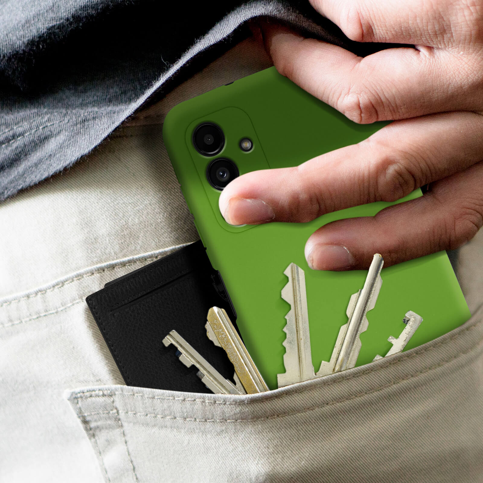 Una mano tiene una custodia verde per telefono in tasca con un portafoglio nero e chiavi, su sfondo chiaro.