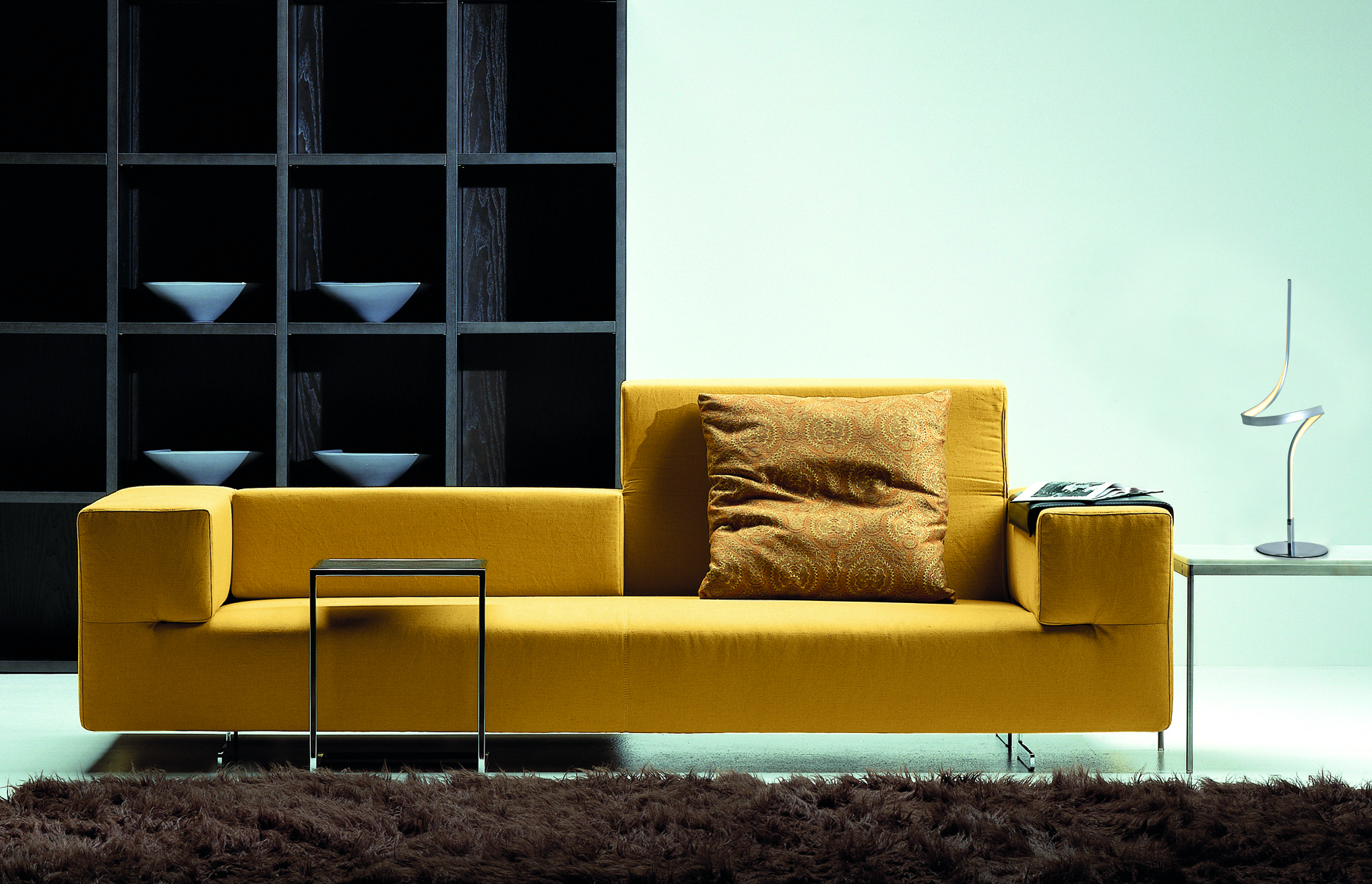 Gelbes Sofa mit braunem Teppich, schwarzem Beistelltisch, Bücherregal im Hintergrund.