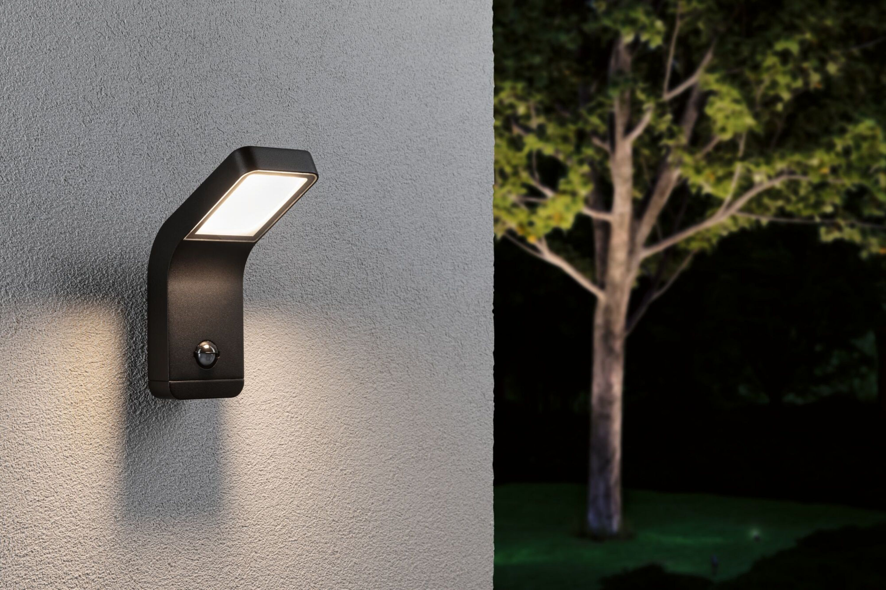 Czarna zewnętrzna lampa ścienna na szarej ścianie, z drzewem w tle.