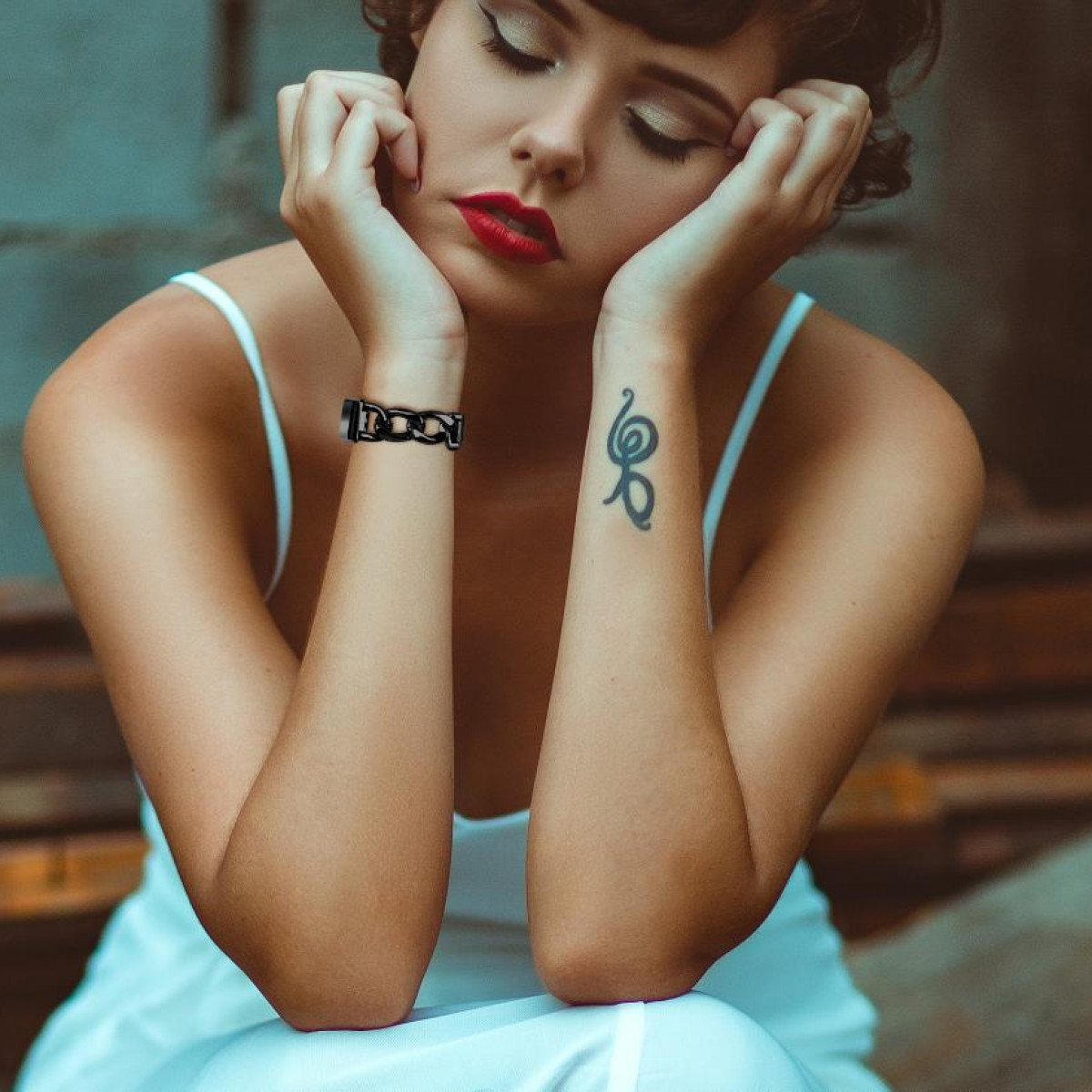 Frau mit rotem Lippenstift und Tattoo, trägt weißes Kleid, stützt Kopf auf Hände, Armband.