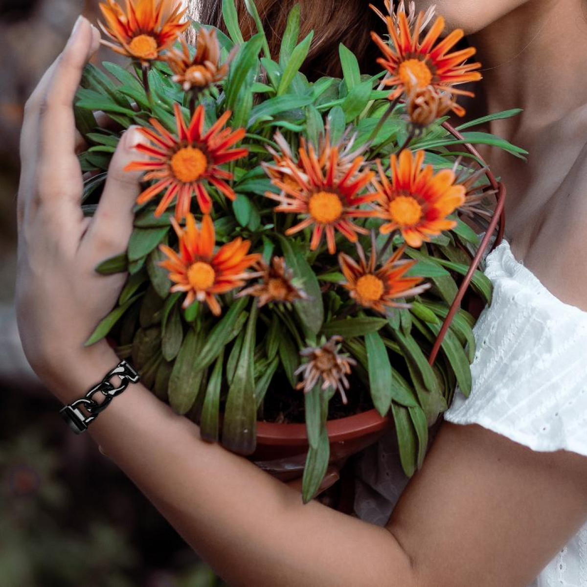 Frau hält eine Topfpflanze mit Gazanien und orangefarbenen Blüten, trägt ein schwarzes Kettenarmband.