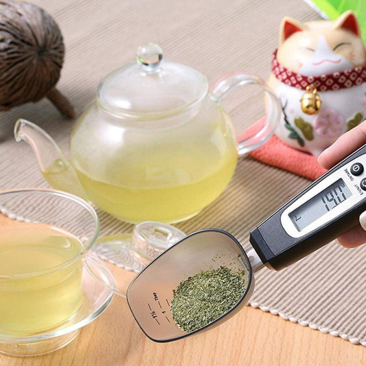 Cyfrowa miarka z zielonymi ziołami. Szklany czajnik i filiżanka w tle.