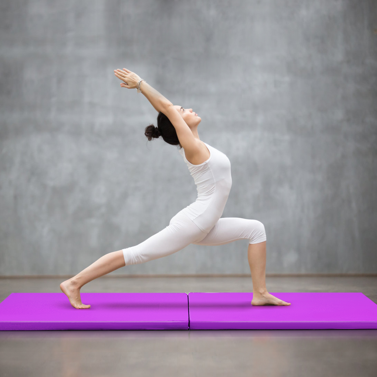 Eine Frau in weißer Yoga-Kleidung in einer Yoga-Pose auf einer lilafarbenen Matte vor einer grauen Wand.