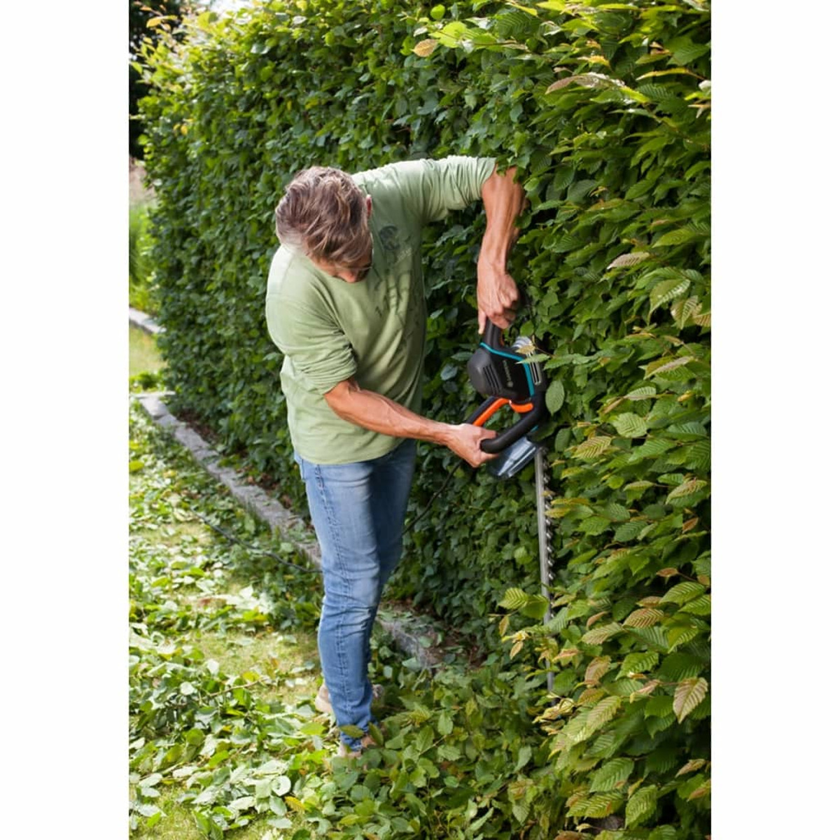 Mann schneidet Hecke mit elektrischem Trimmer, trägt grünes Hemd, sonniger Tag.