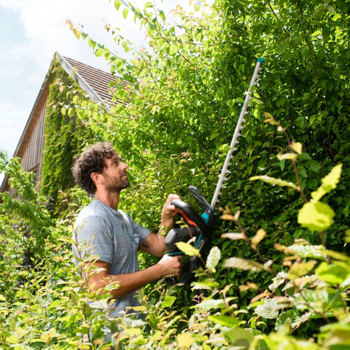 Ein Mann schneidet eine grüne Hecke mit einem elektrischen Trimmer. Im Hintergrund sind ein Holzgebäude und Himmel.