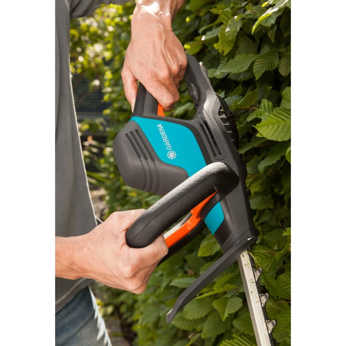 Person benutzt eine schwarz-blaue Heckenschere mit orangen Akzenten. Der Hintergrund ist grünes Laub.