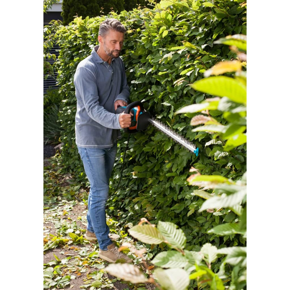 Ein Mann schneidet eine Hecke mit einem elektrischen Heckenschneider. Grüne Hecke und Pflanzen im Hintergrund.