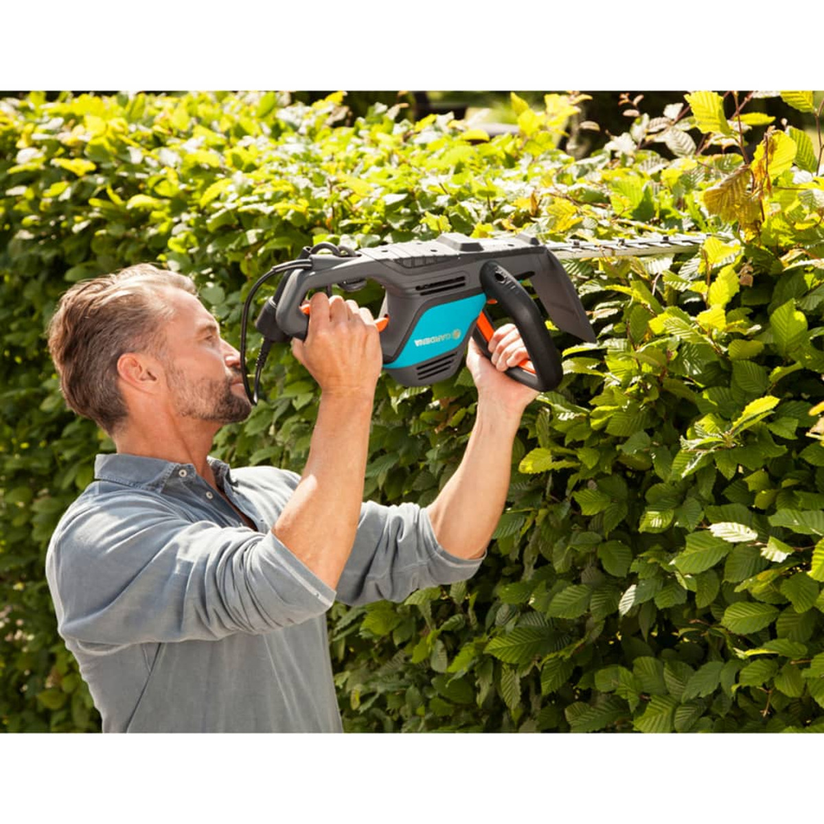 Ein Mann schneidet eine grüne Hecke mit einem elektrischen Trimmer. Der Hintergrund ist grünes Laub.