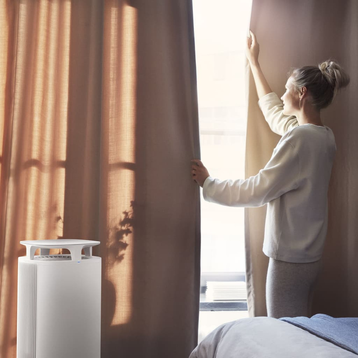 Frau öffnet braune Vorhänge, links ein weißer Luftreiniger. Hintergrund: Fenster, Bett.
