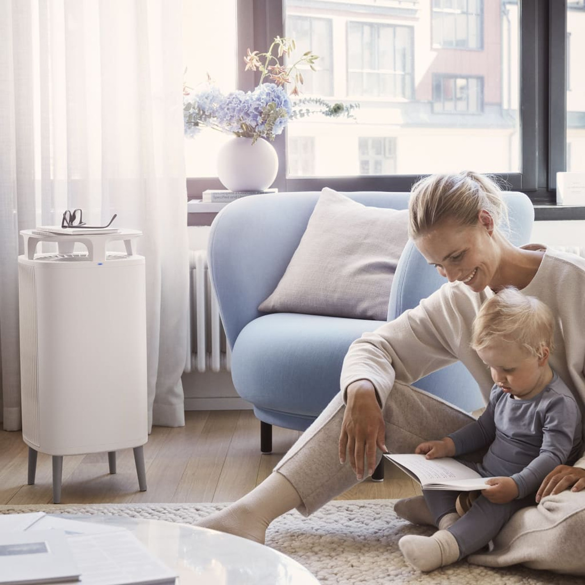 Eine Frau und ein Kind sitzen in einem Wohnzimmer. Es gibt einen Luftreiniger und eine Blumenvase.