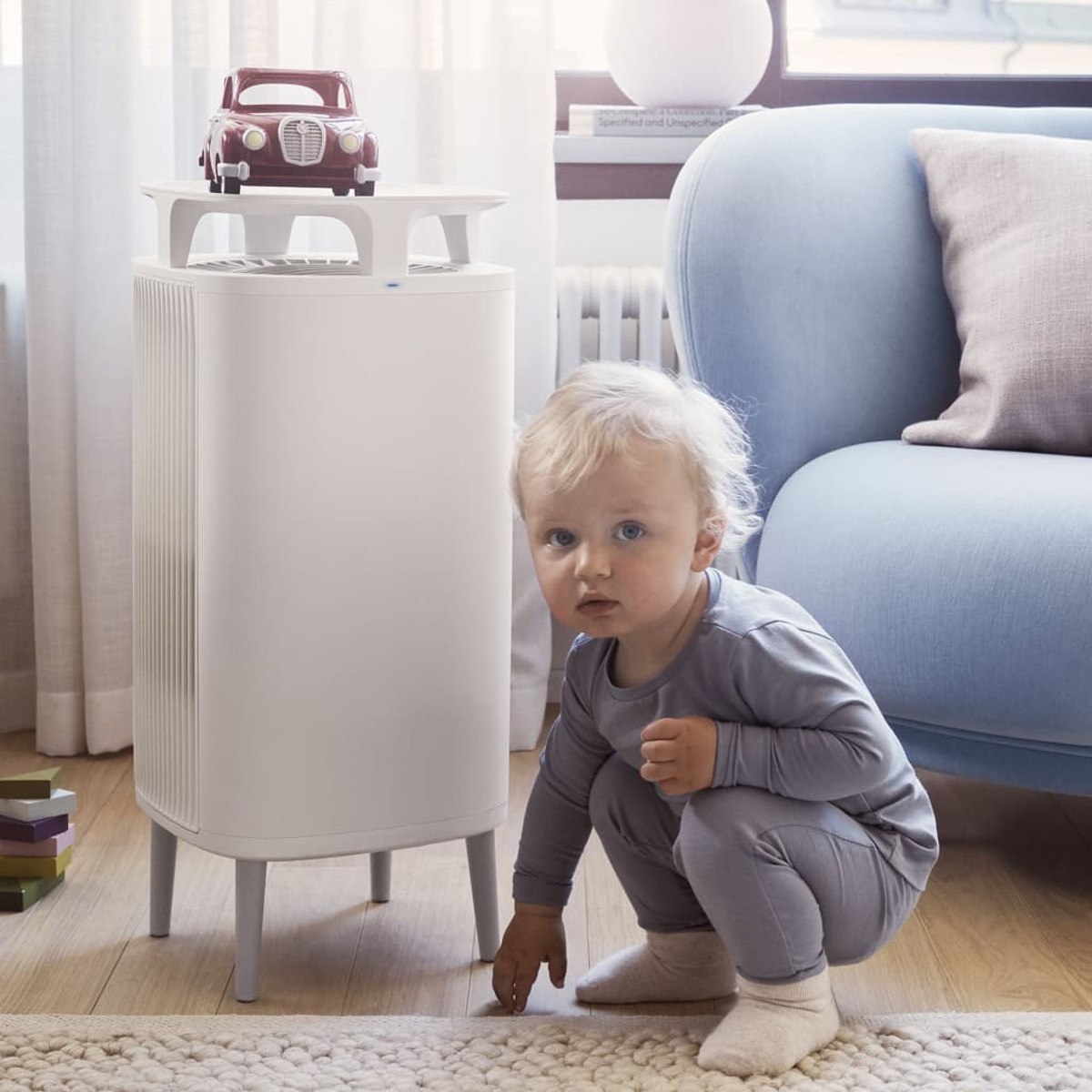 Ein Baby hockt neben einem Luftreiniger. Ein Spielzeugauto steht oben drauf. Eine Couch ist im Hintergrund.