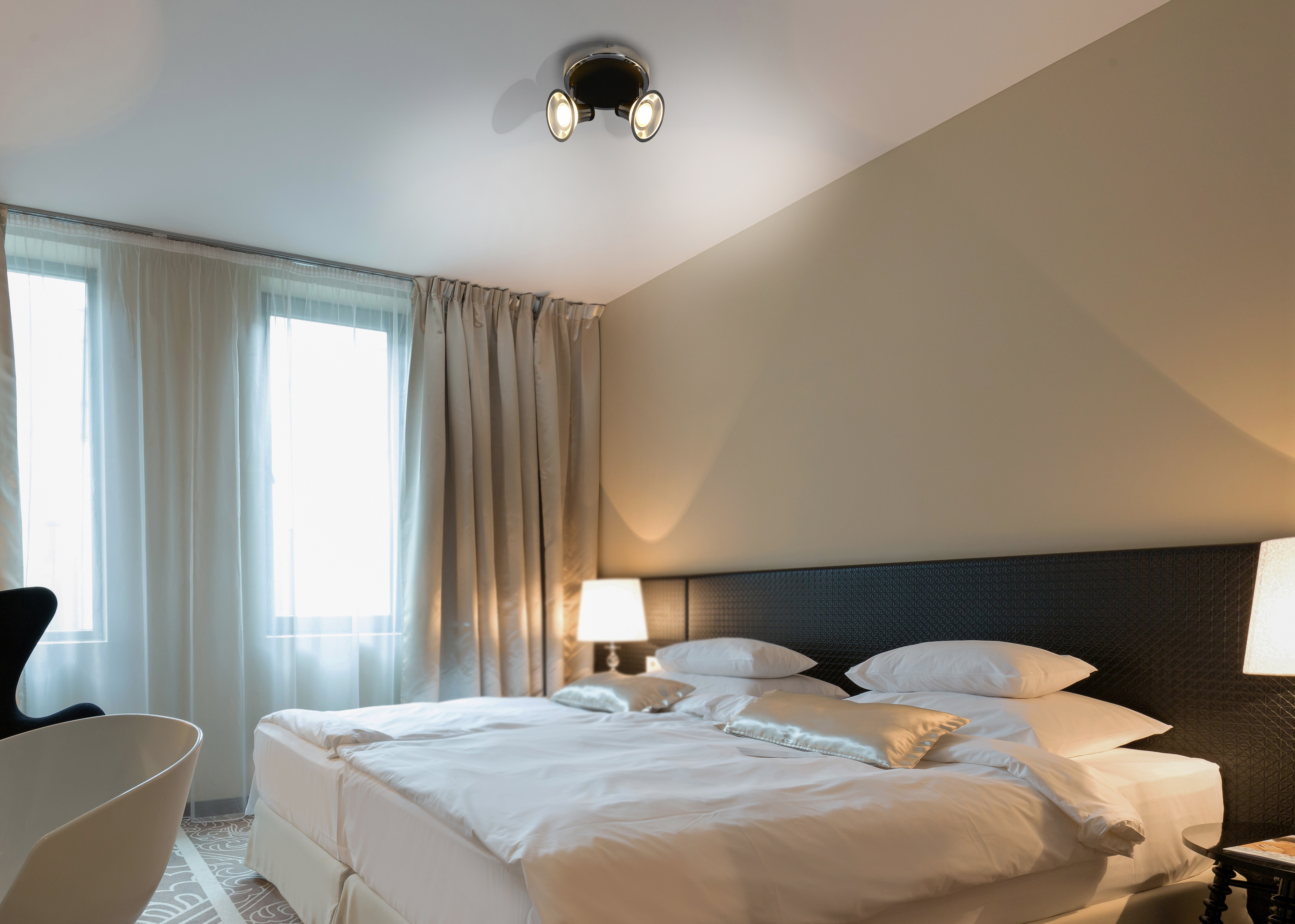 Ein Schlafzimmer mit Bett, Lampen und Deckenleuchte. Es gibt Fenster und Vorhänge.