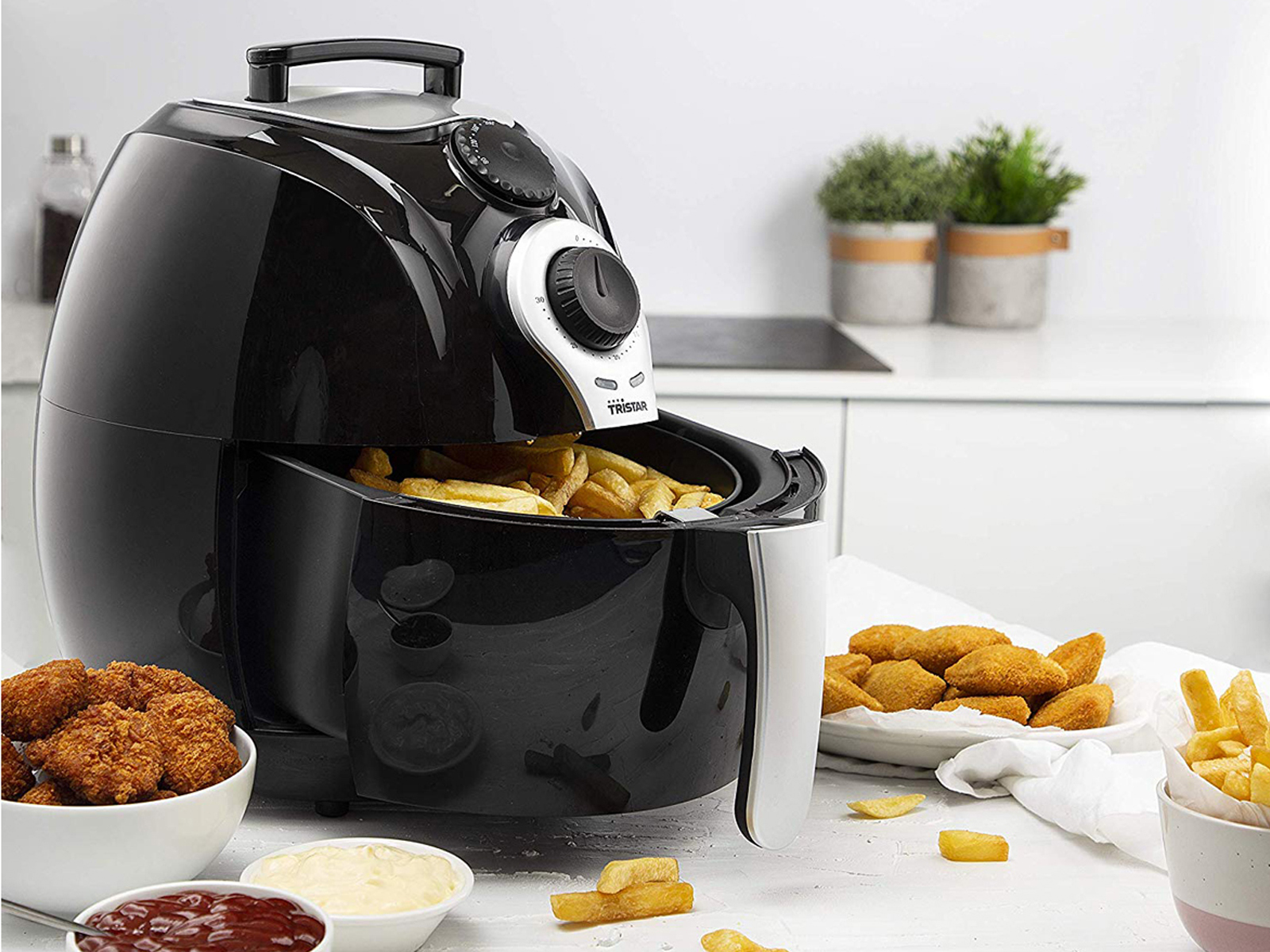 Freidora de aire negra con patatas fritas dentro. Comida frita como nuggets y patatas fritas en superficie blanca.