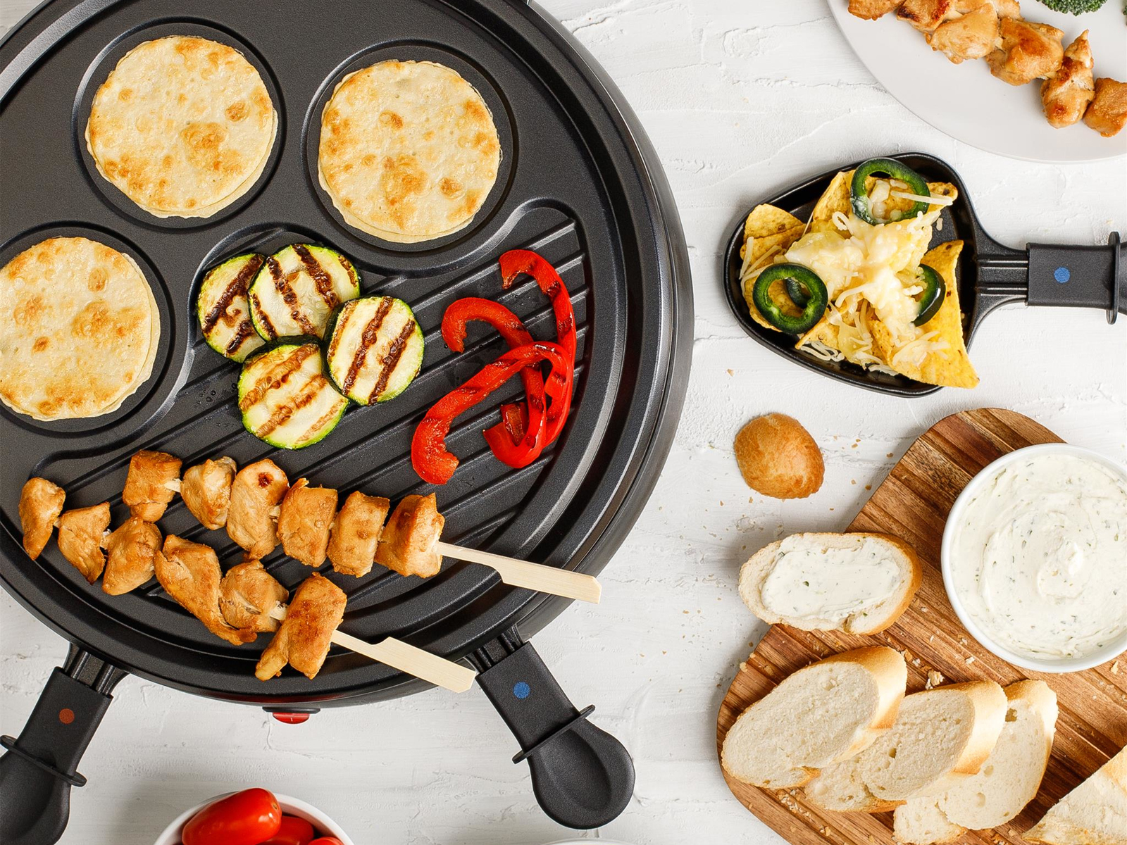 Ein Raclette-Grill mit Essen: Pfannkuchen, gegrilltes Gemüse, Hähnchenspieße, Nachos, Brot und Dip.