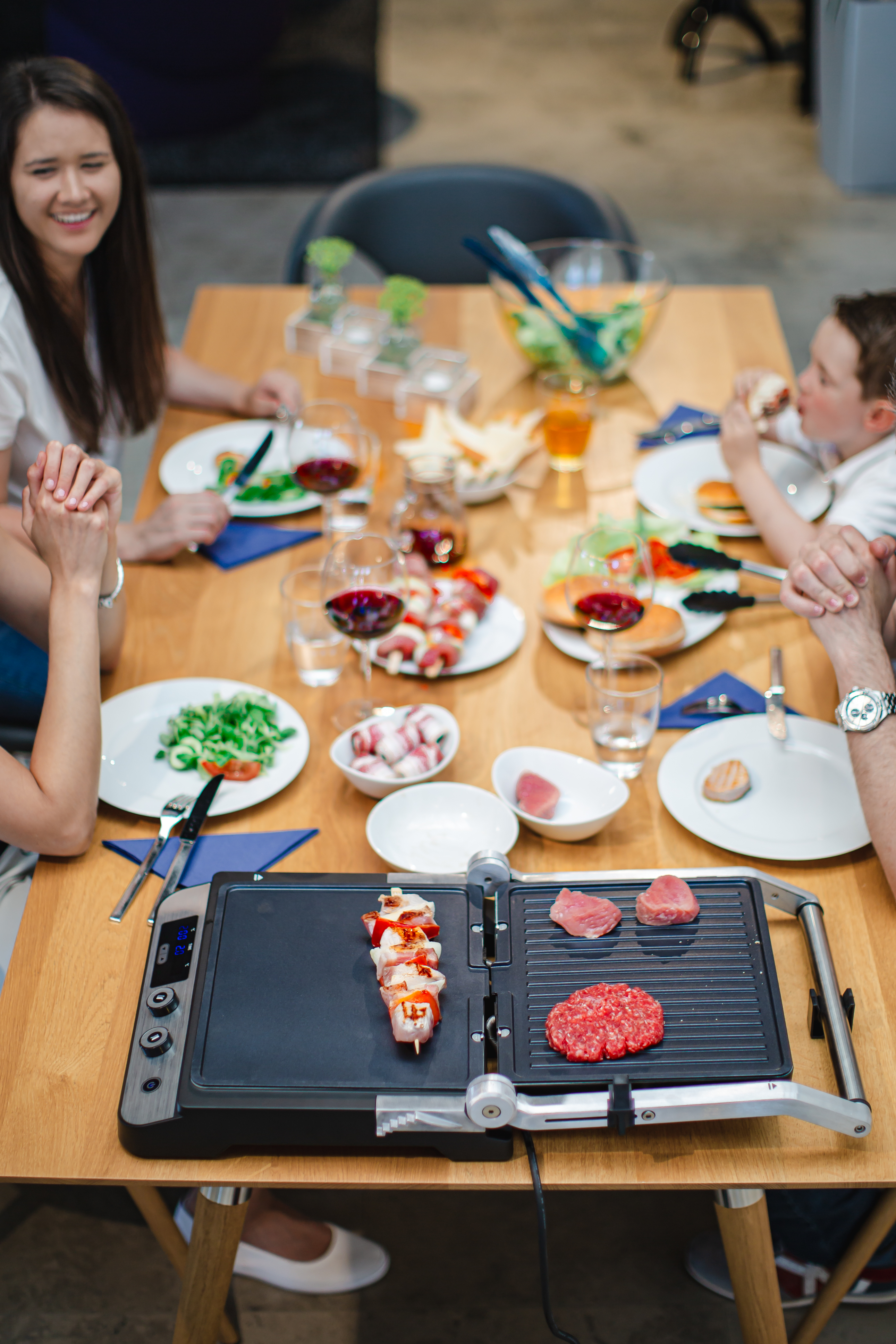 Menschen um einen Holztisch mit Essen und einem Grill. Spieße und Burger werden gegrillt.