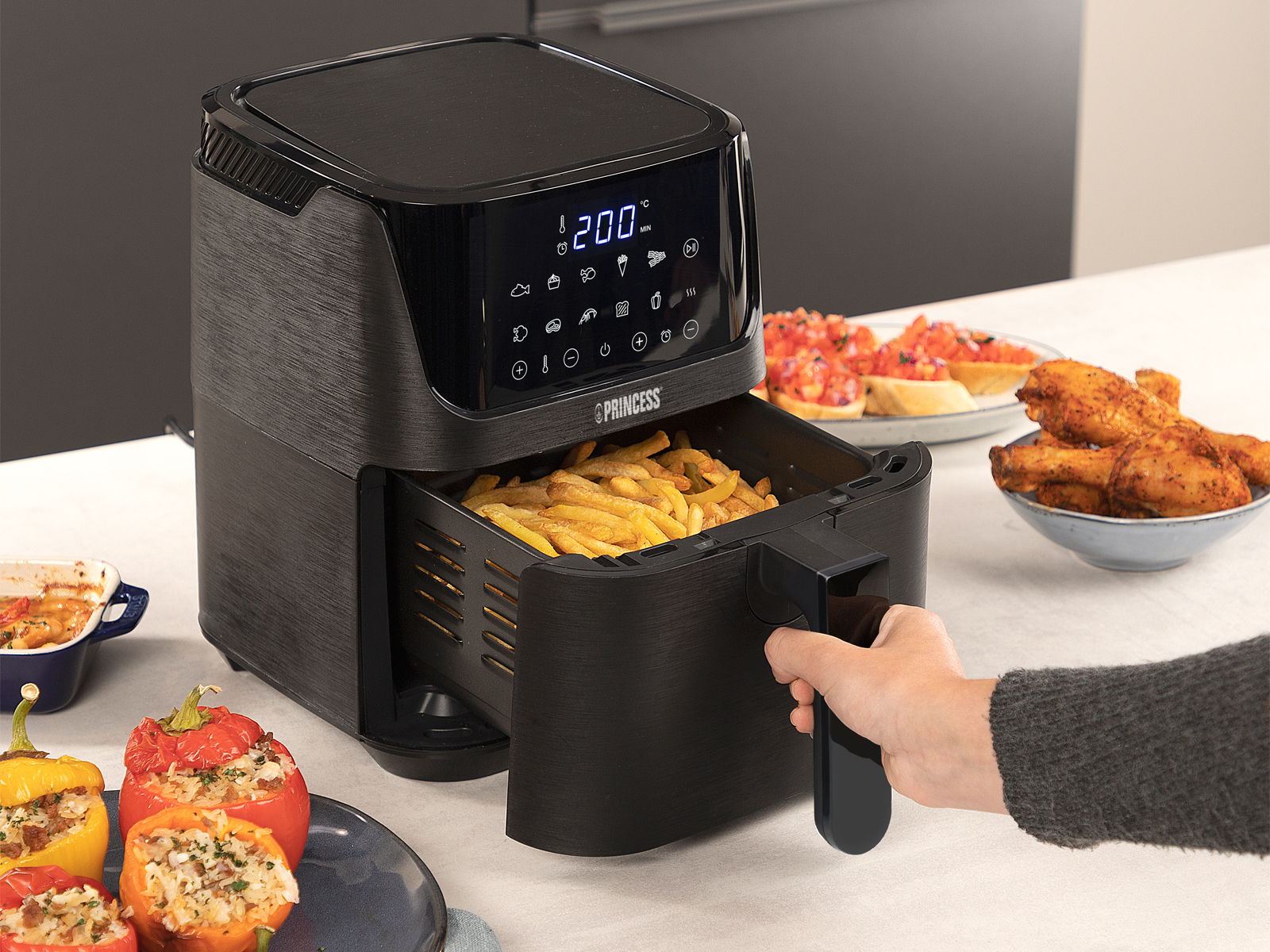 Freidora de aire negra con pantalla digital, con patatas fritas en la cesta. Hay alimentos como pimientos en el fondo.