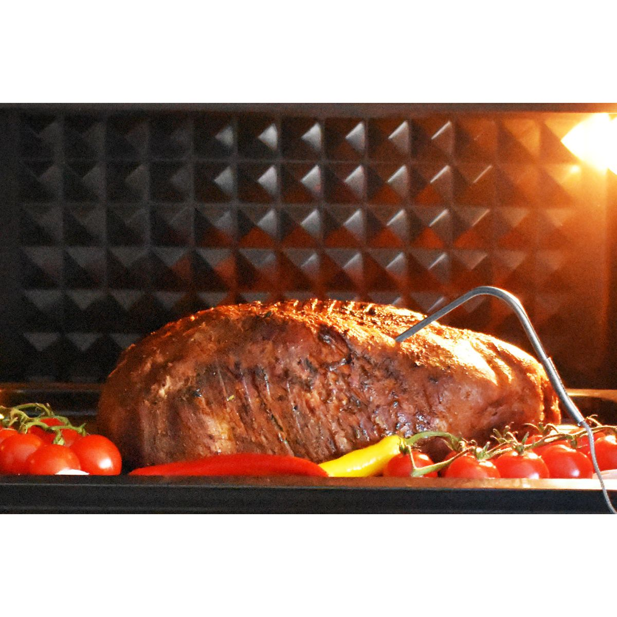 Ein gebratener Hackbraten im Ofen, mit Tomaten und Paprika. Eine Temperatursonde ist in das Fleisch eingeführt.