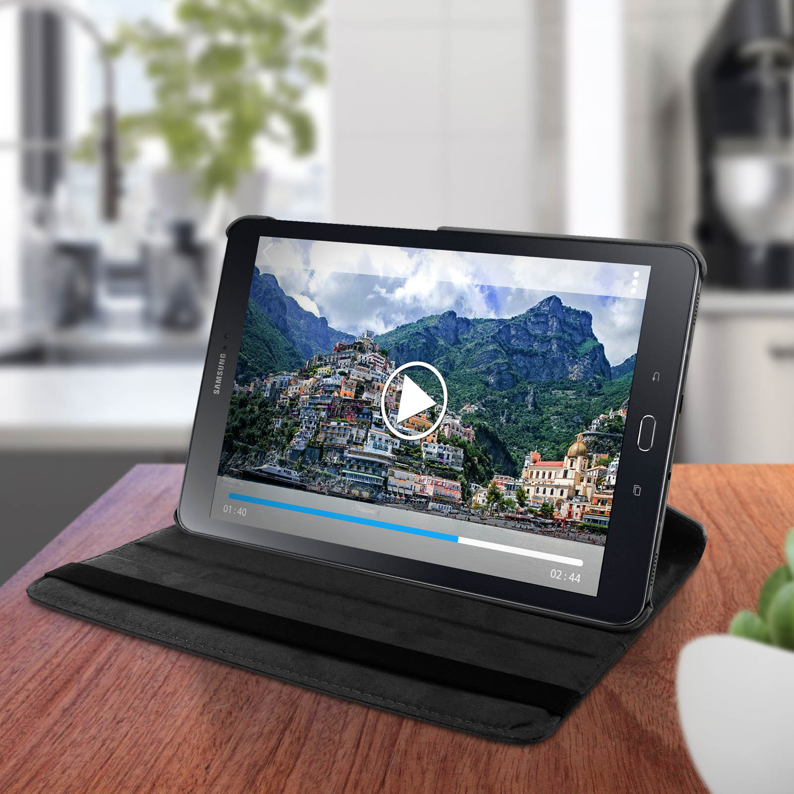 Tablet su un tavolo di legno, che riproduce un video. Uno sfondo mostra una città costiera ed elementi della cucina.