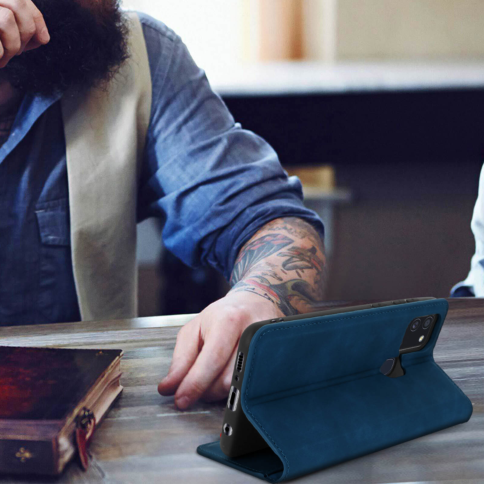 Un uomo con la barba usa un telefono in una custodia di pelle blu, su un tavolo.