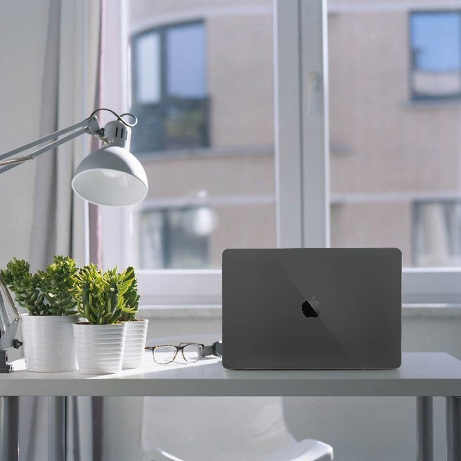 Laptop auf weißem Schreibtisch in Fensternähe. Schreibtischlampe und Pflanzen sichtbar.