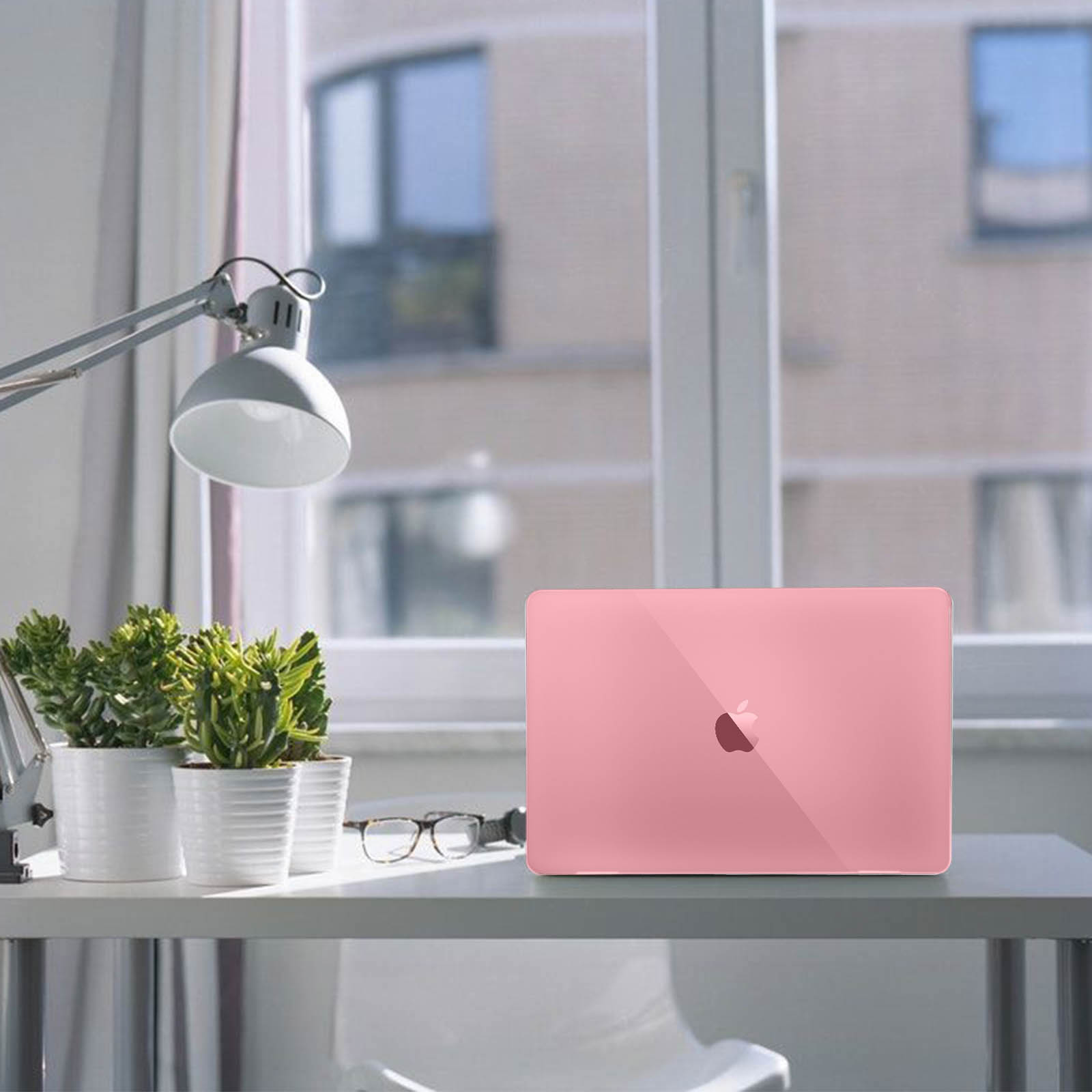 Ein pinkfarbenes Laptop auf einem weißen Schreibtisch mit Pflanzen, einer Lampe und Brille.