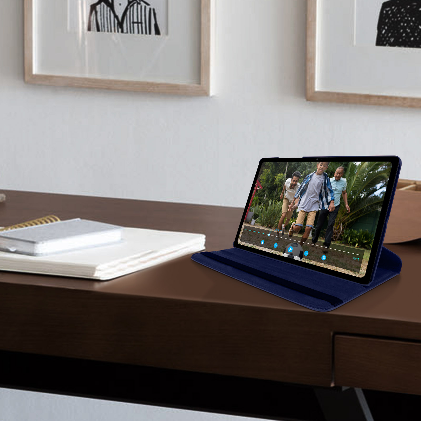 Tablet mostra un video, appoggiato sulla scrivania. Custodia blu aperta. Sfondo: scrivania in legno, due quadri.