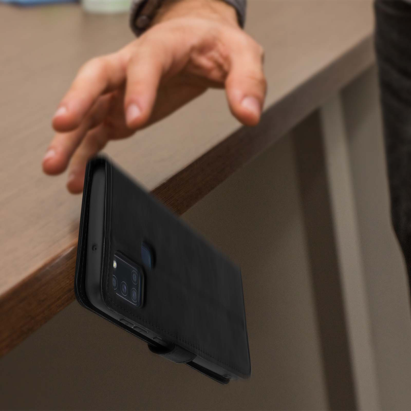 Een hand reikt naar een zwarte telefoonhoes aan de rand van een bruine tafel.
