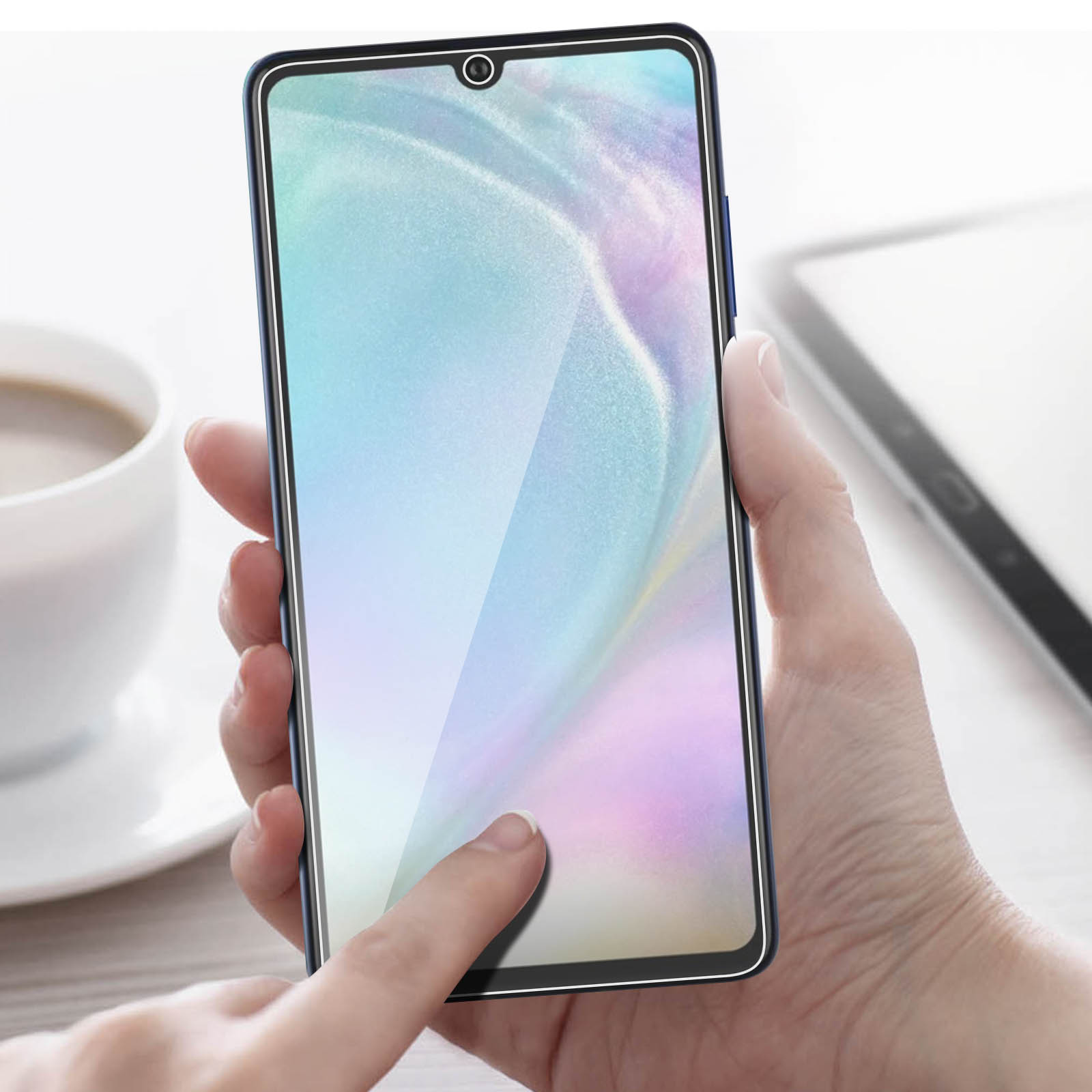 Una mano tocca lo schermo di un telefono con una protezione per lo schermo. Una tazza e un tablet sono sullo sfondo.