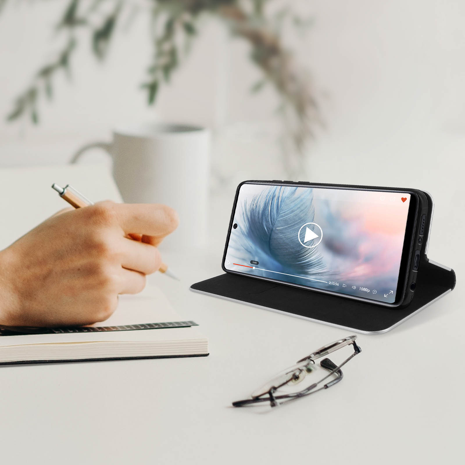 Telefono su un supporto che riproduce un video, occhiali e quaderno su una scrivania.