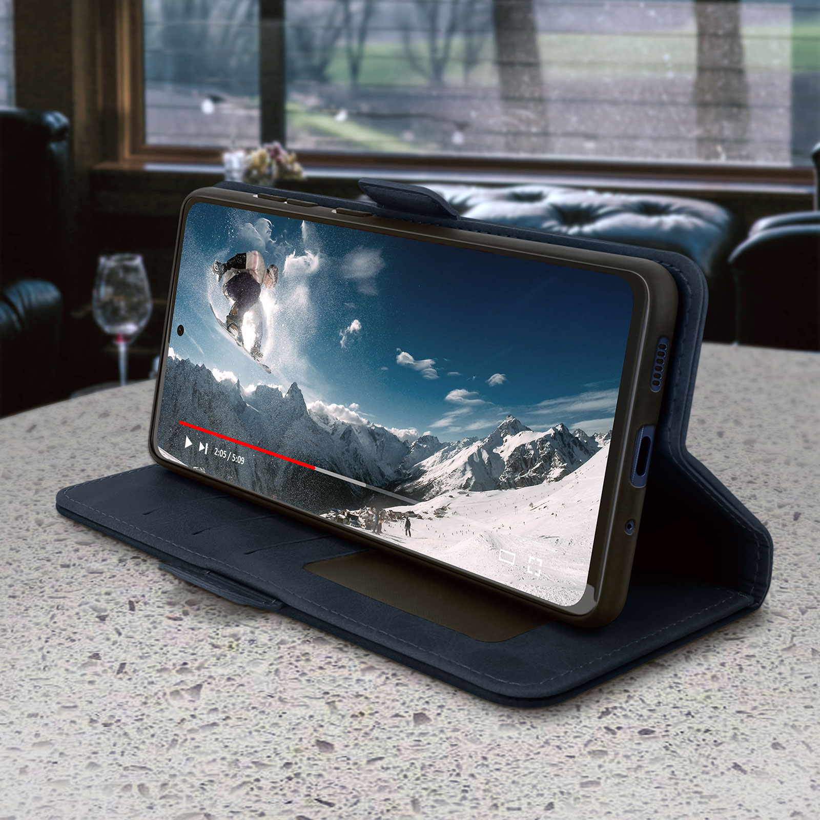Téléphone affichant une vidéo de skieur sur une table. Étui en cuir bleu. Fond : paysage montagneux.