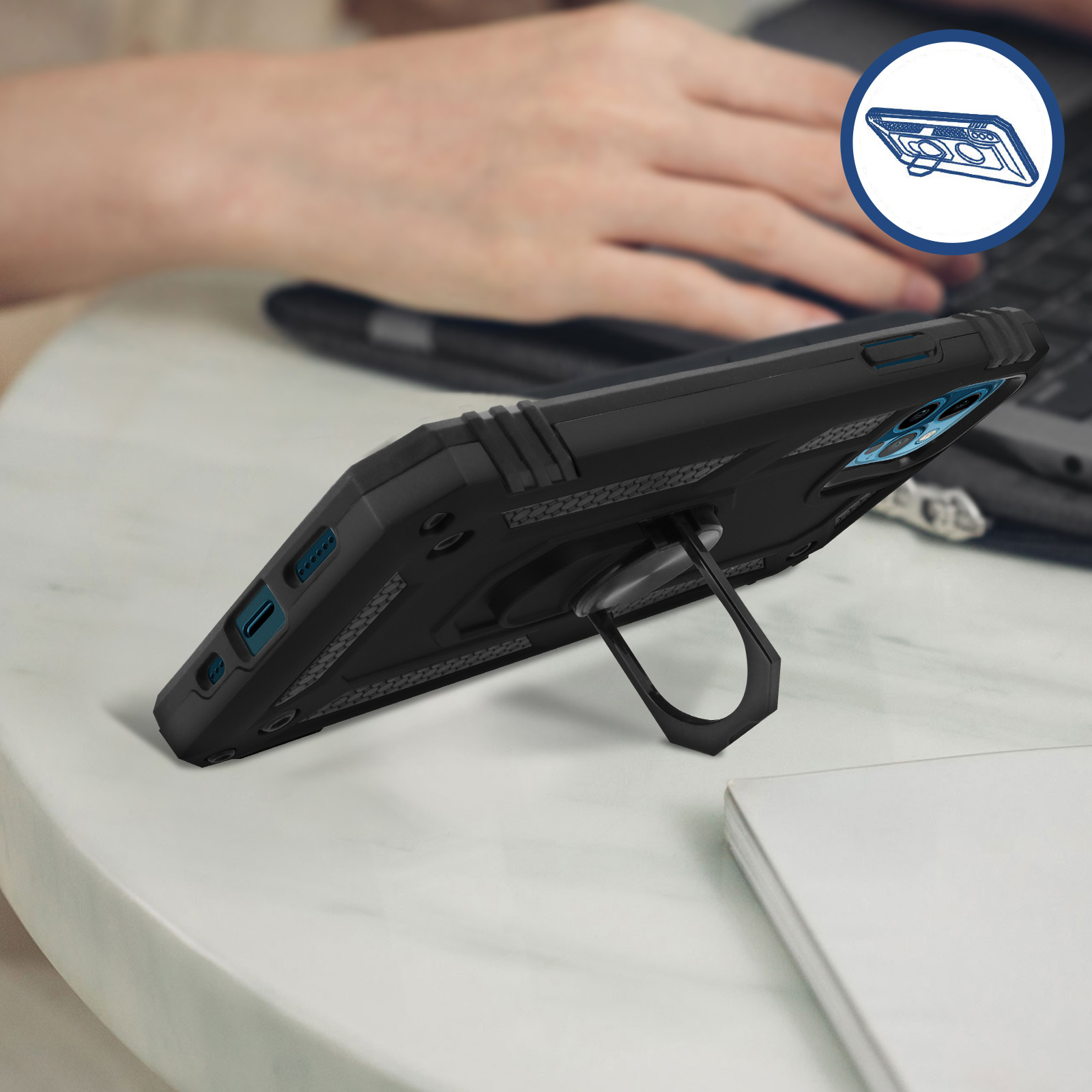 Una custodia per telefono nera con un supporto ad anello. Una persona usa un laptop. Un tavolo bianco in primo piano.