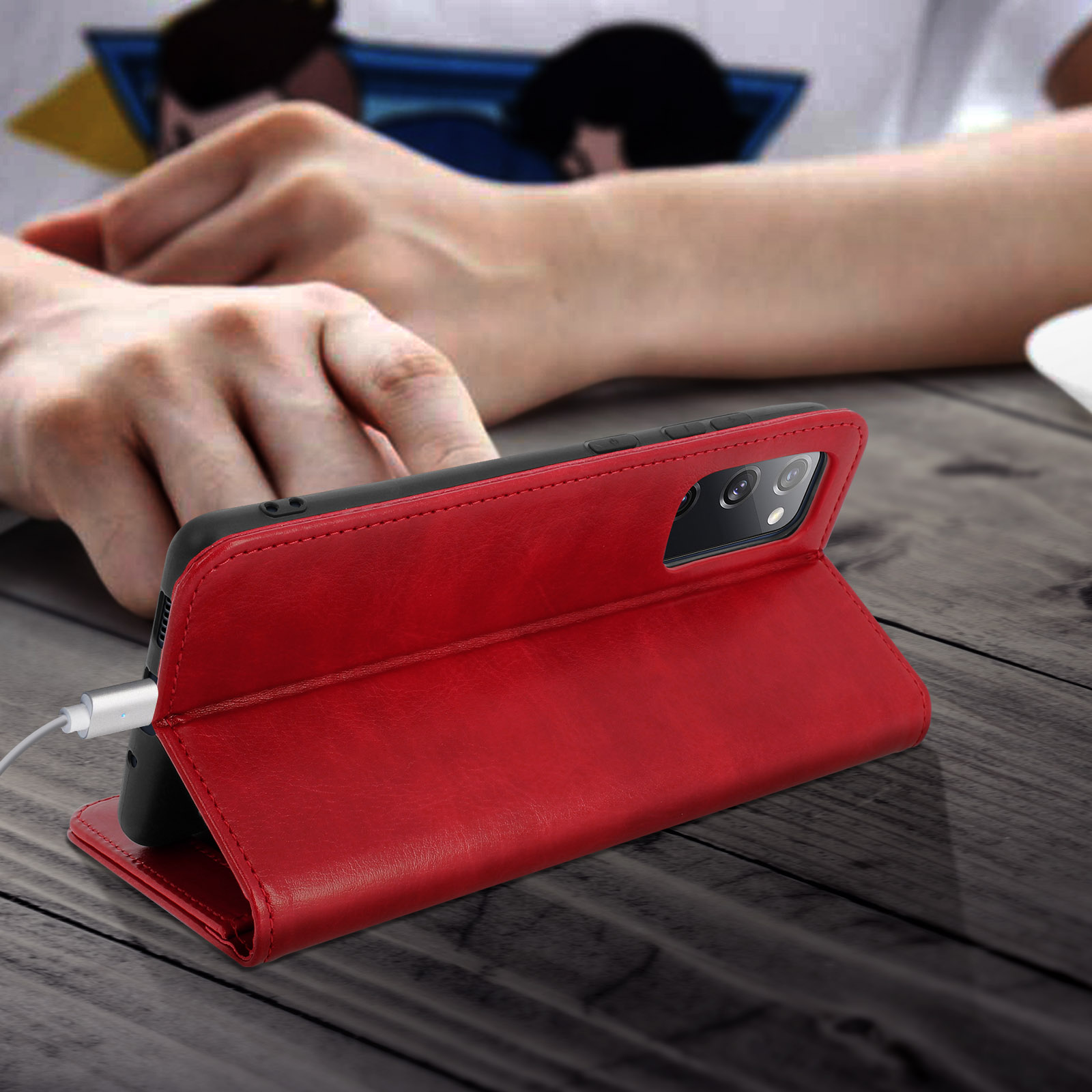 Een telefoon in een rode hoes leunt tegen een houten tafel, verbonden met een oplaadkabel.