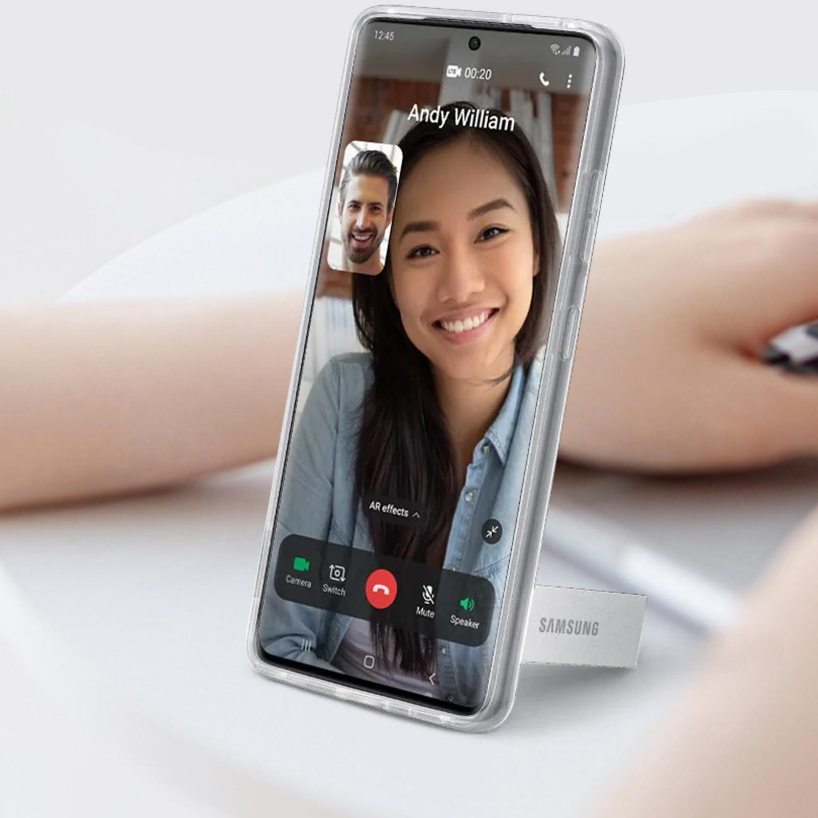 Un telefono mostra una videochiamata. Una donna sorride. Il telefono è in una custodia trasparente. Uno sfondo è visibile.
