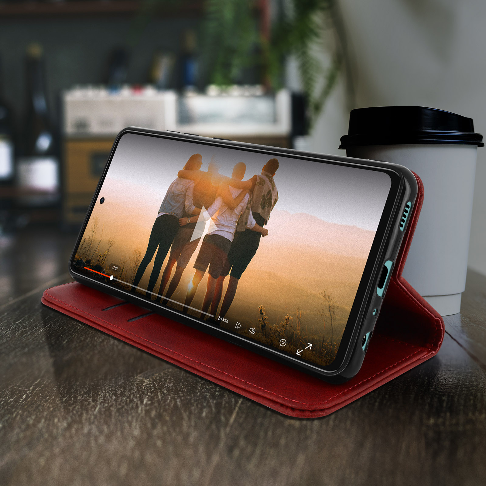 Telefono in custodia rossa mostra video di persone che si abbracciano, su tavolo in legno con tazza.