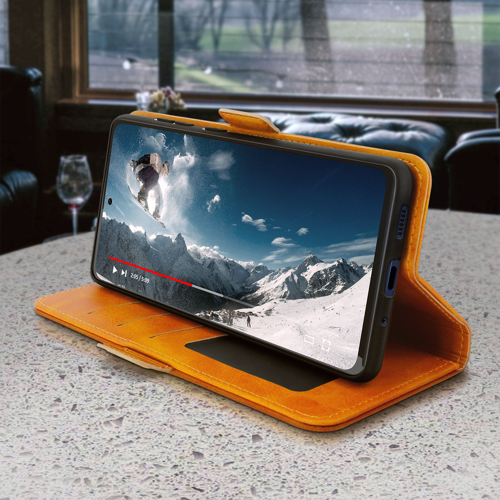 Téléphone dans un étui orange ouvert, affichant une vidéo d'un snowboarder sur une table dans une pièce.