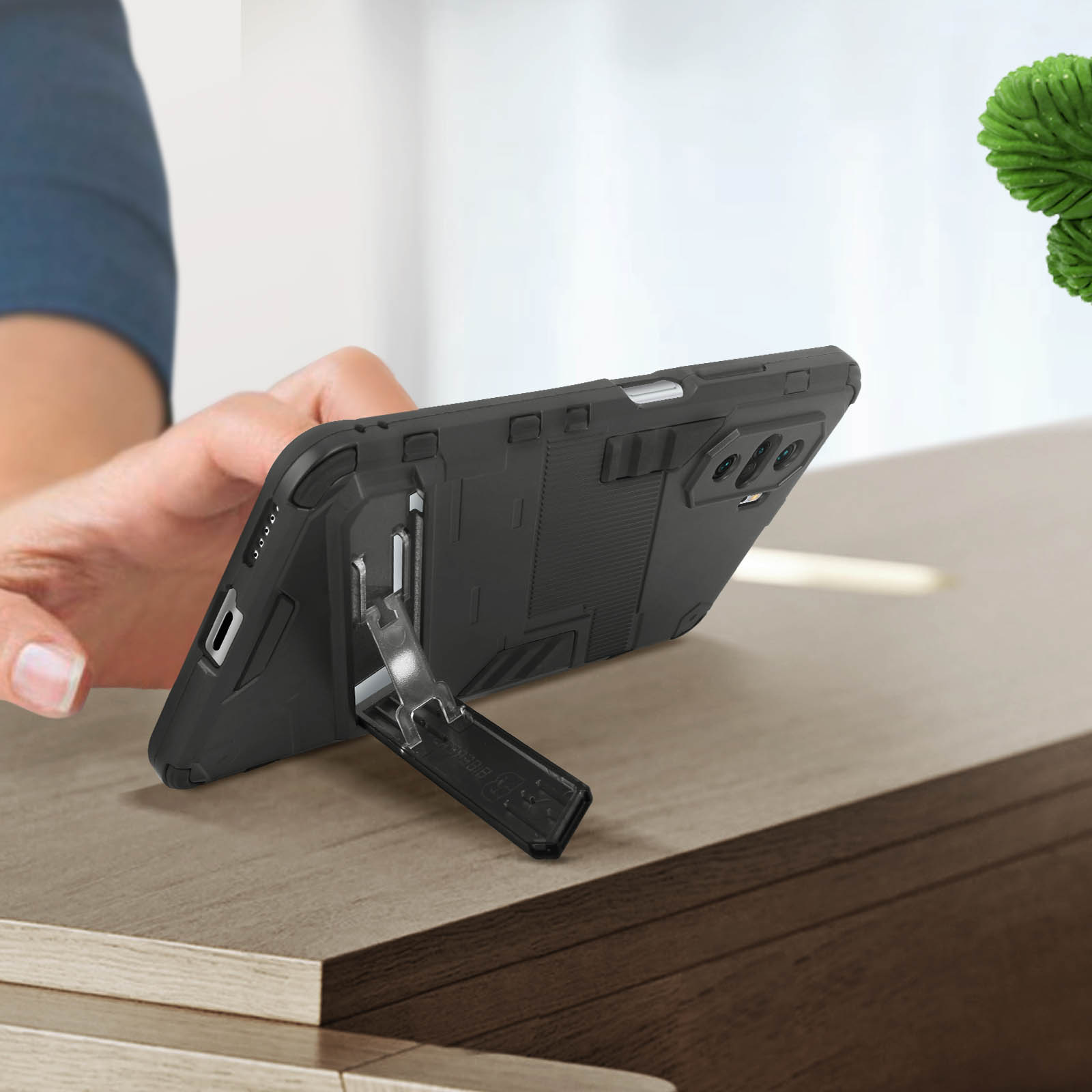Custodia per telefono nera con cavalletto, su una superficie di legno. Una mano tocca il telefono.