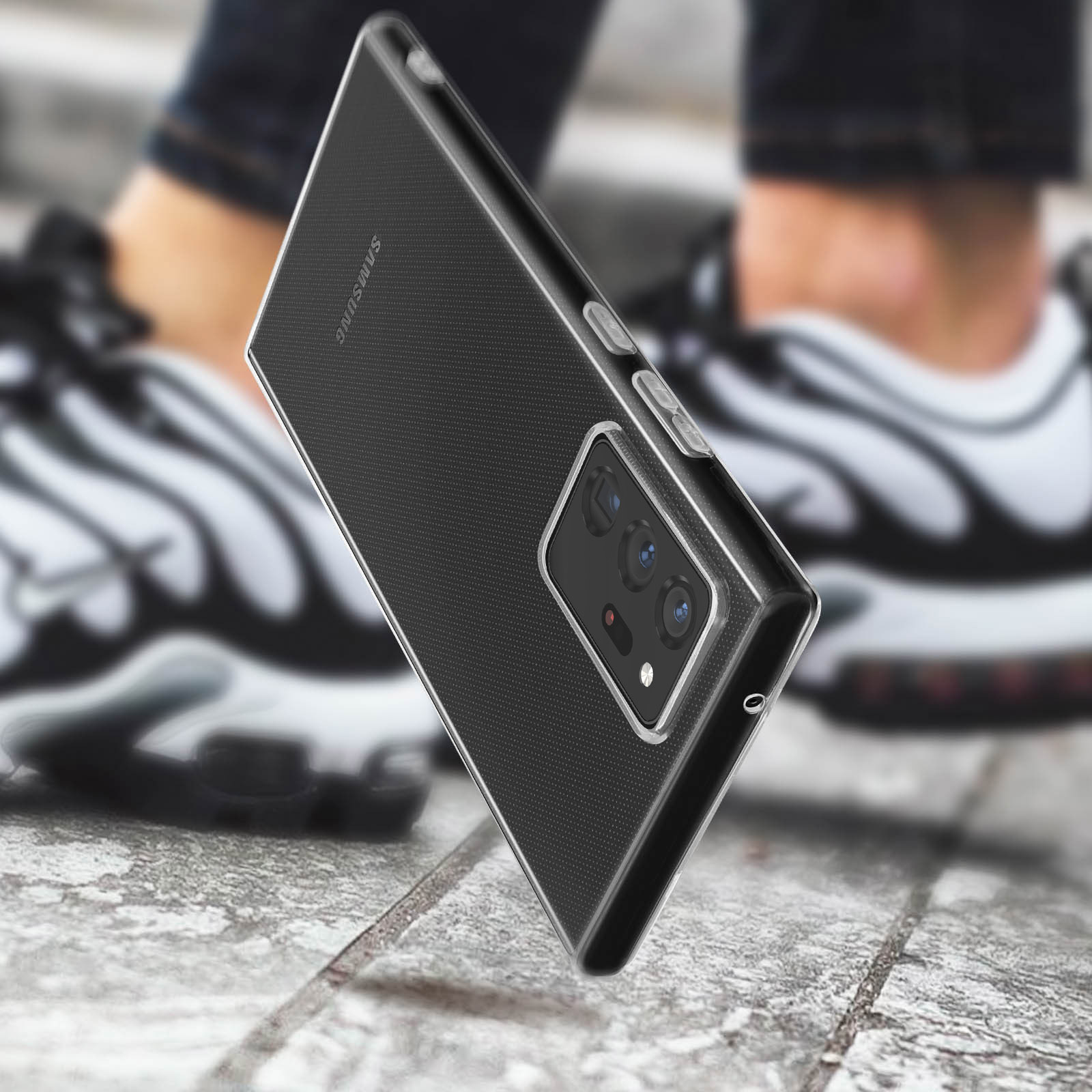 Een telefoonhoesje met een zwarte, getextureerde achterkant wordt in de buurt van een paar wazige sneakers gehouden.