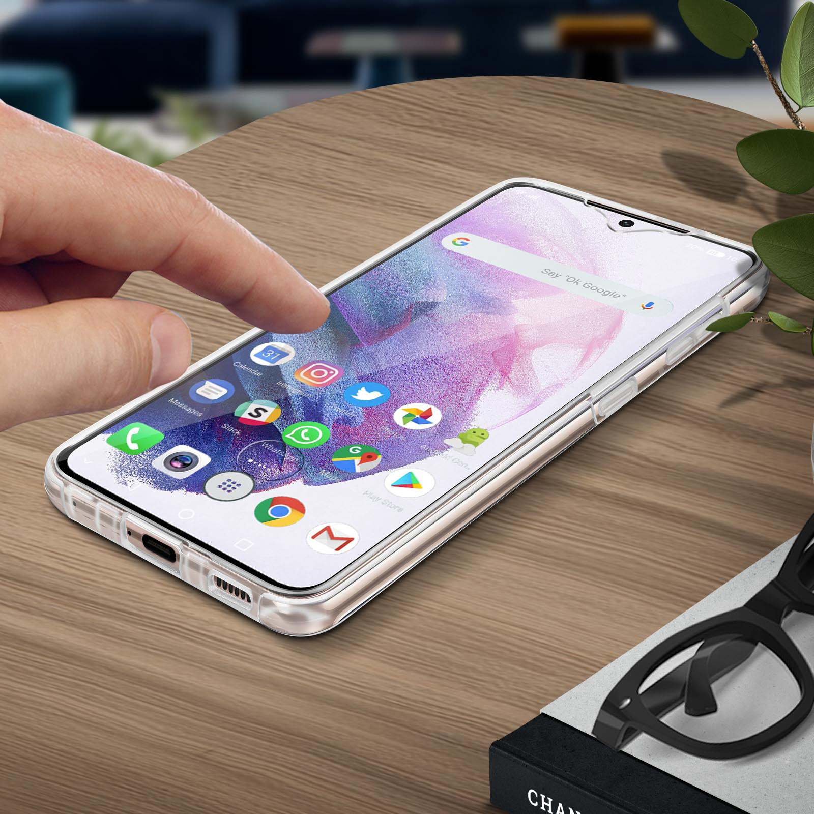 Una mano tocca lo schermo di un telefono su un tavolo. Le app sono visibili. Pianta e occhiali sullo sfondo.