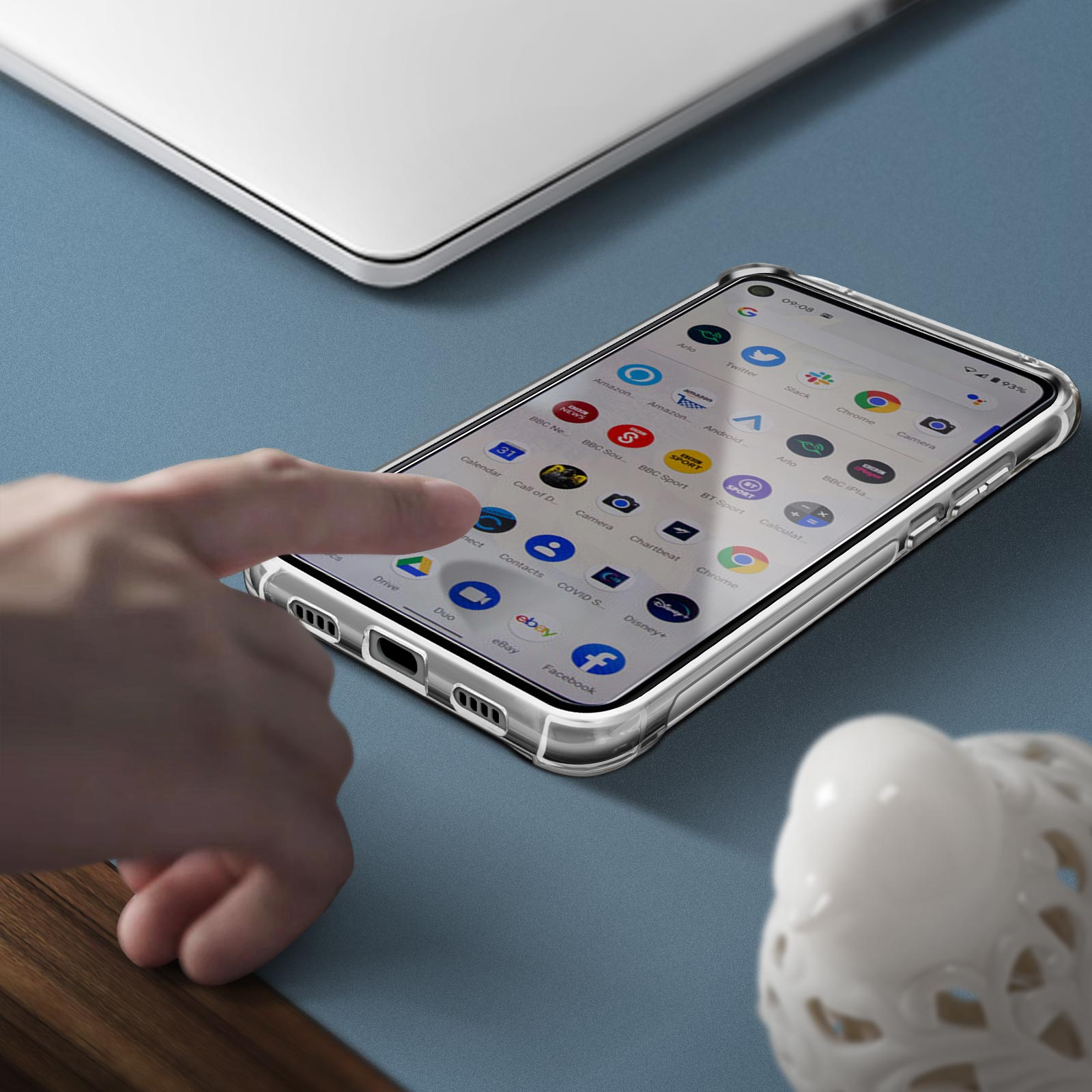 Mano tocca un telefono in una custodia trasparente su superficie blu, laptop e scultura bianca vicini.