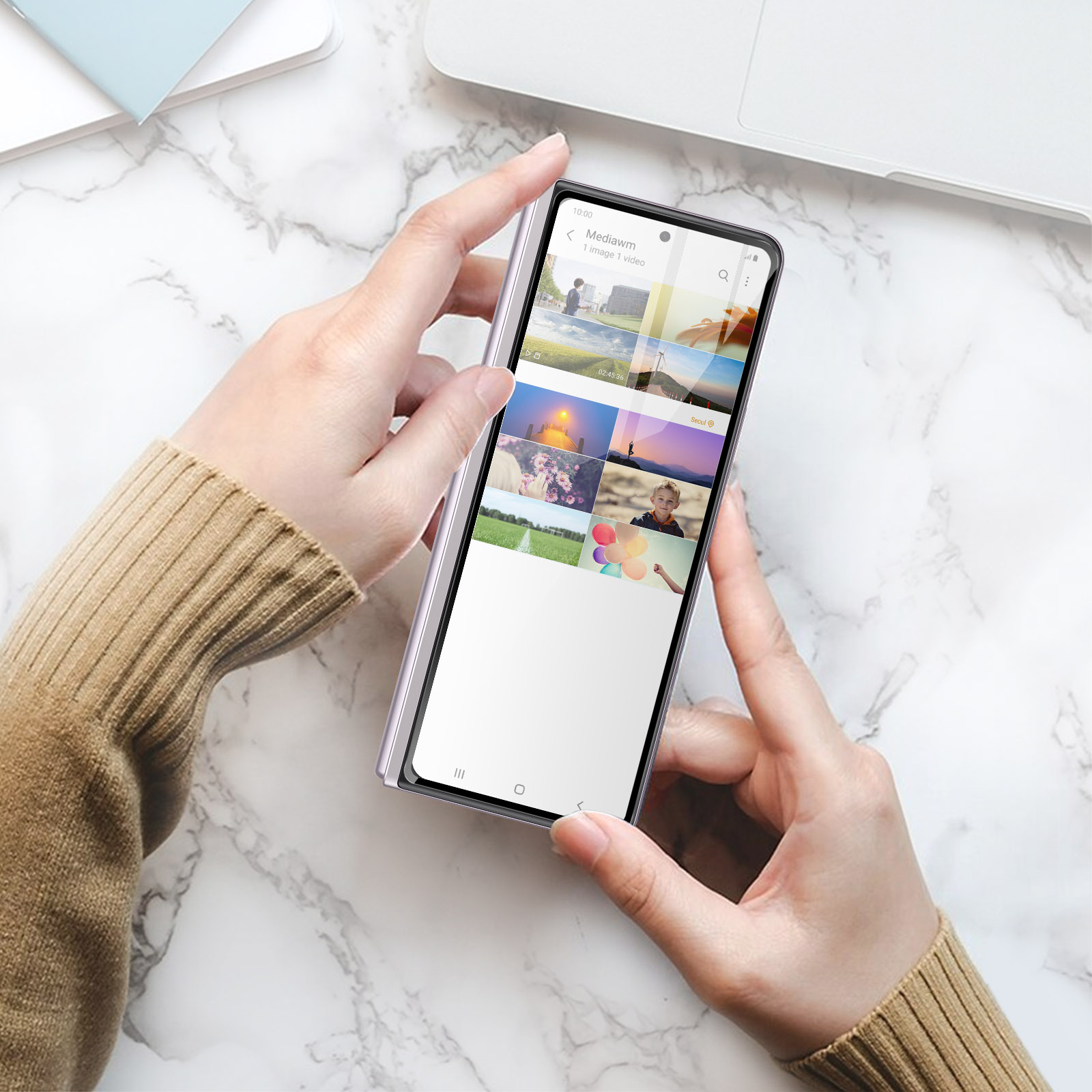 Mani che tengono un telefono che mostra miniature di foto. Il telefono è su una superficie di marmo con un laptop e fogli sullo sfondo.