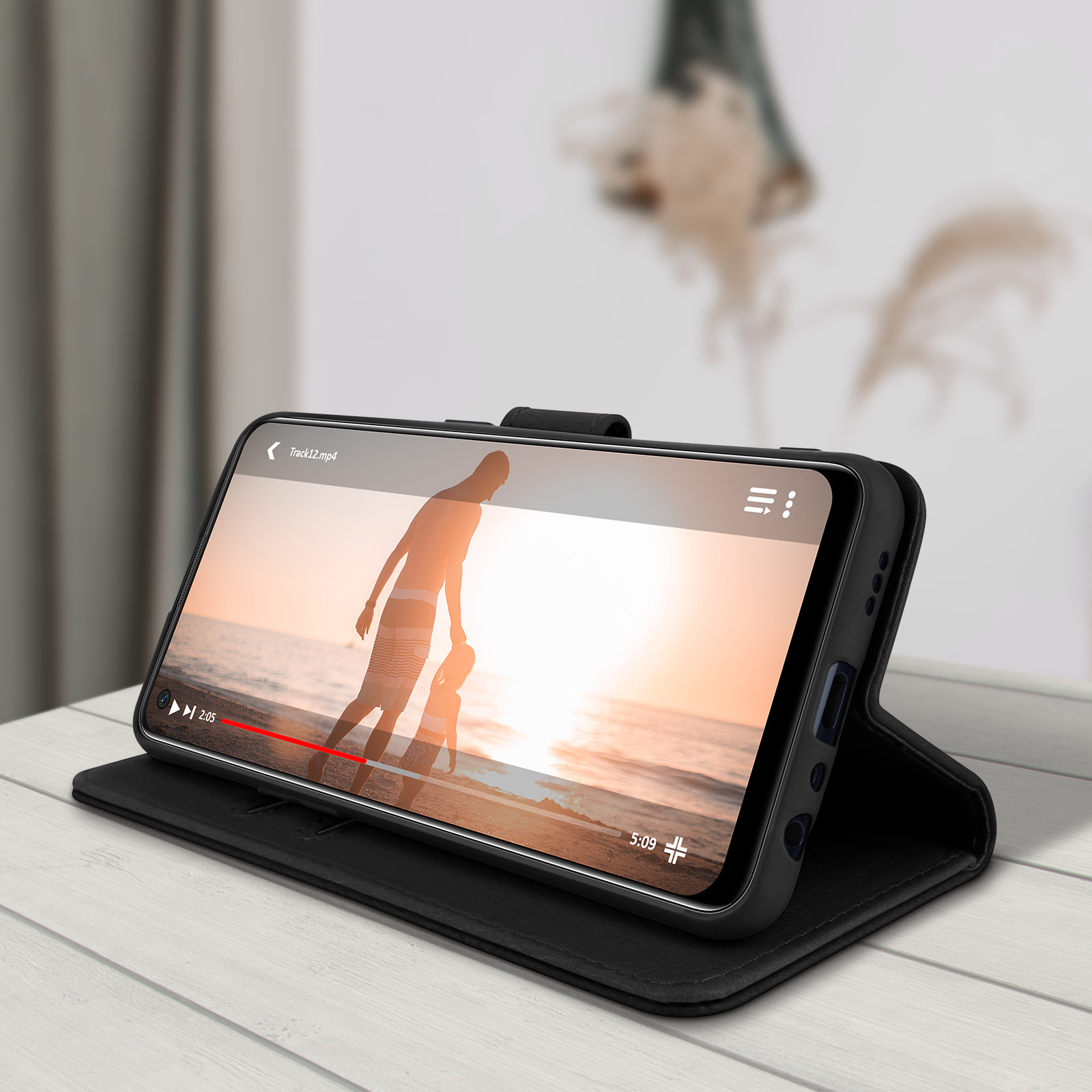 Un étui de téléphone noir contient un téléphone affichant une vidéo d'un couple sur une plage. Il est posé sur une table en bois.