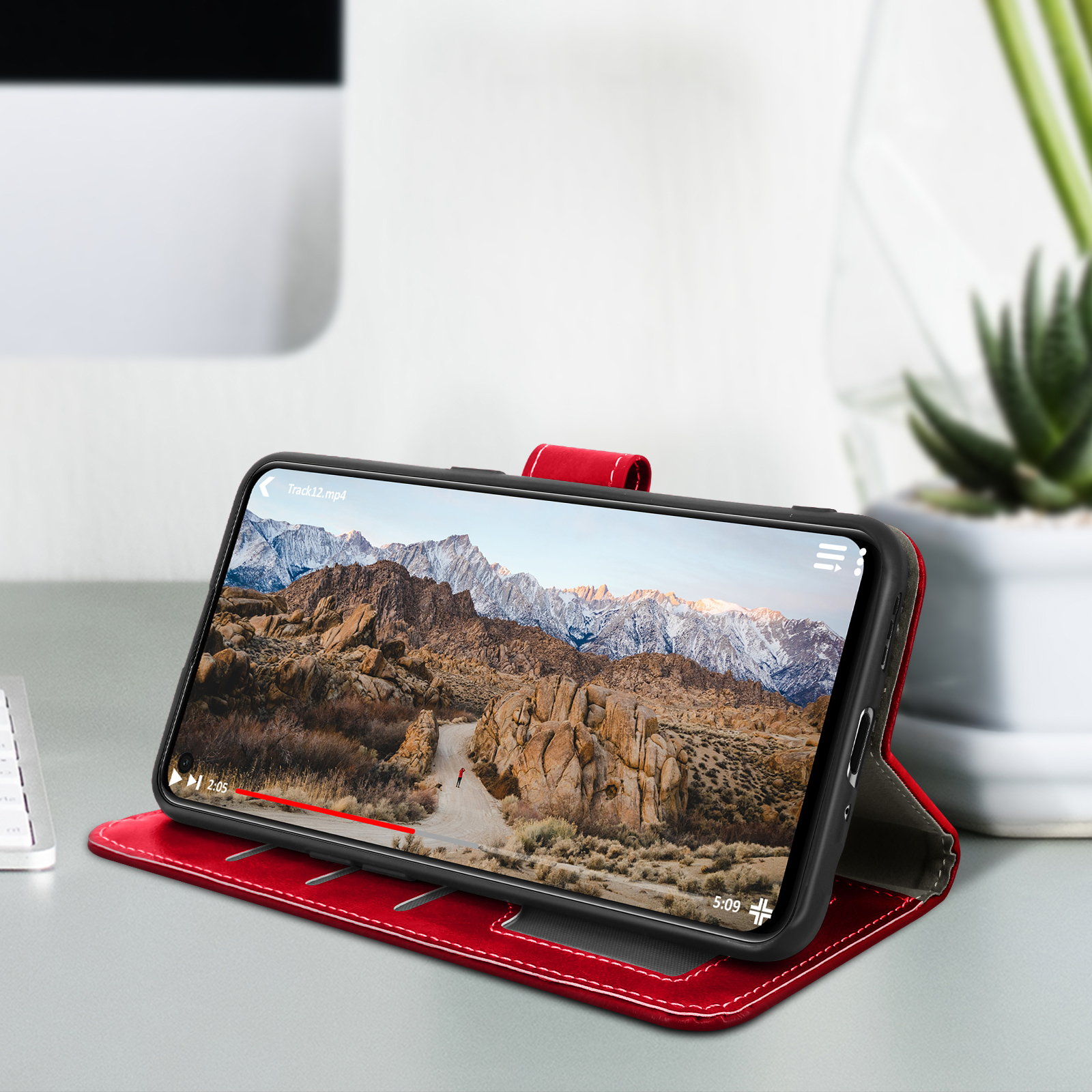 Téléphone affichant une vidéo de paysage, dans un étui rouge. Le fond est un bureau blanc et une plante.