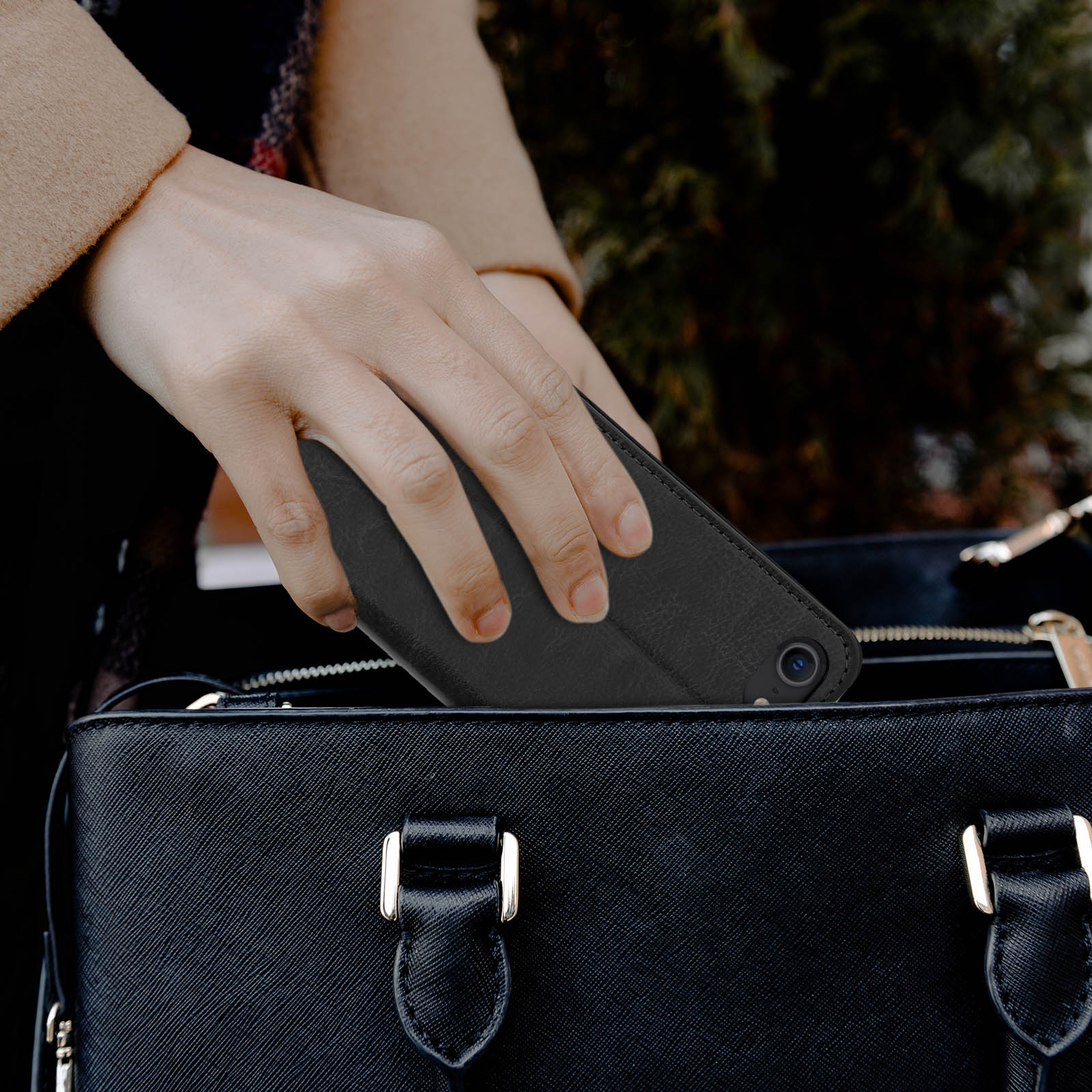 Una mano mette un telefono nero in una borsa nera. La borsa è su uno sfondo chiaro.