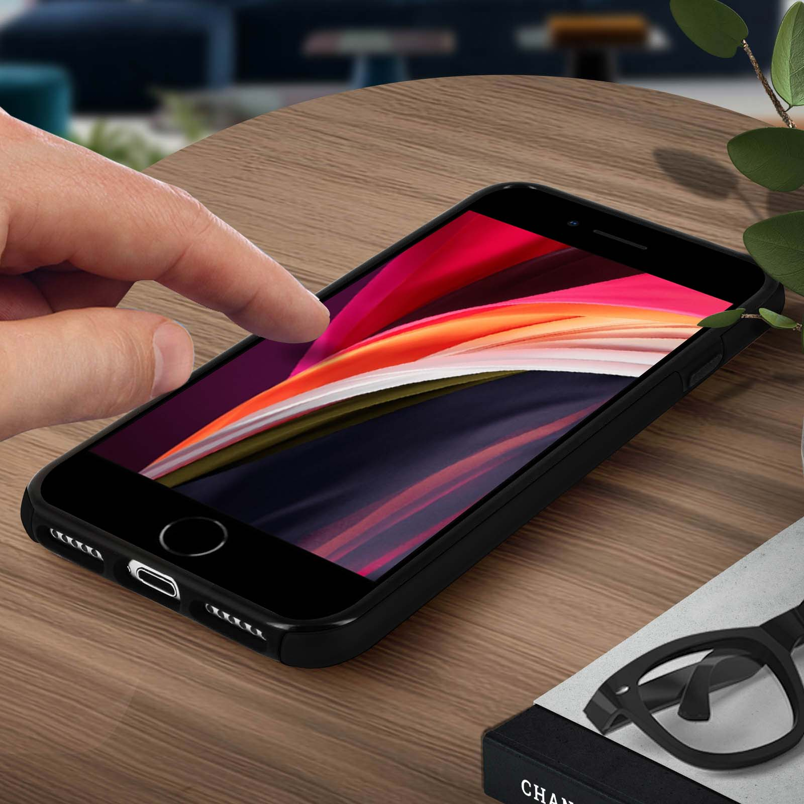 Una mano tocca lo schermo di un telefono su un tavolo di legno, occhiali e libro accanto.