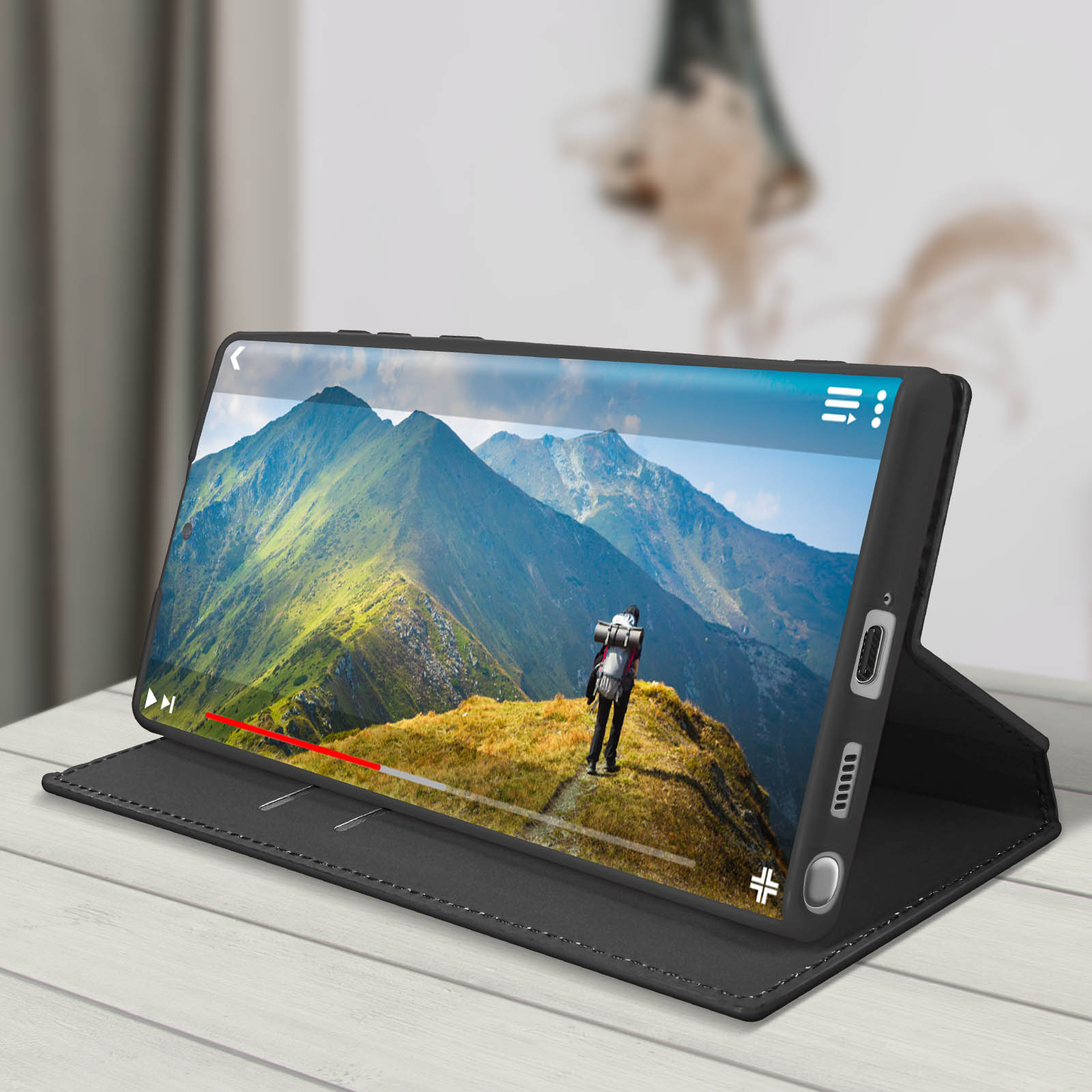 Un téléphone dans un étui noir est ouvert, montrant une vidéo de paysage de montagne. Il repose sur une surface blanche.