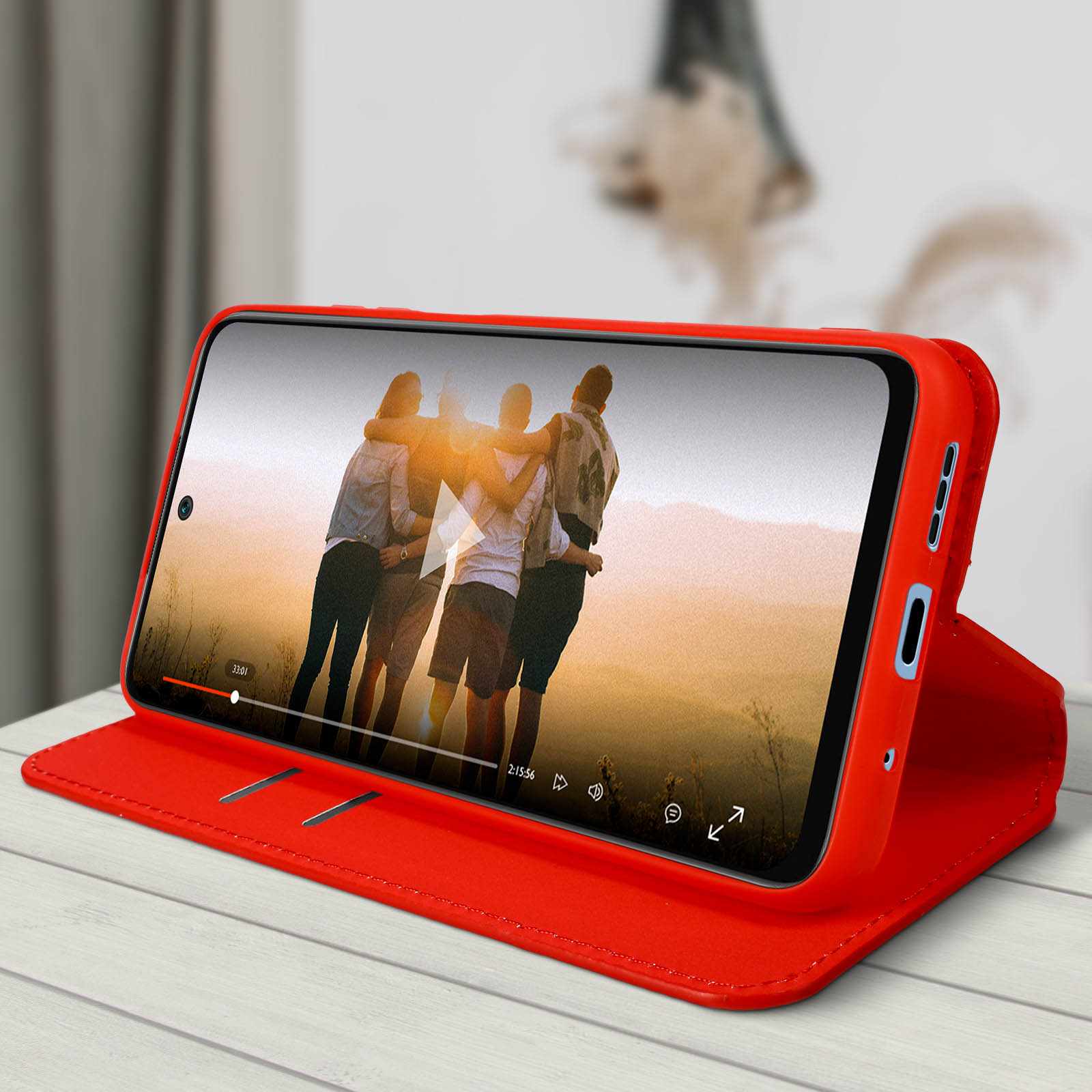Una custodia per telefono rossa con un telefono all'interno è aperta, mostrando un video di persone. L'ambientazione è su un tavolo di legno.