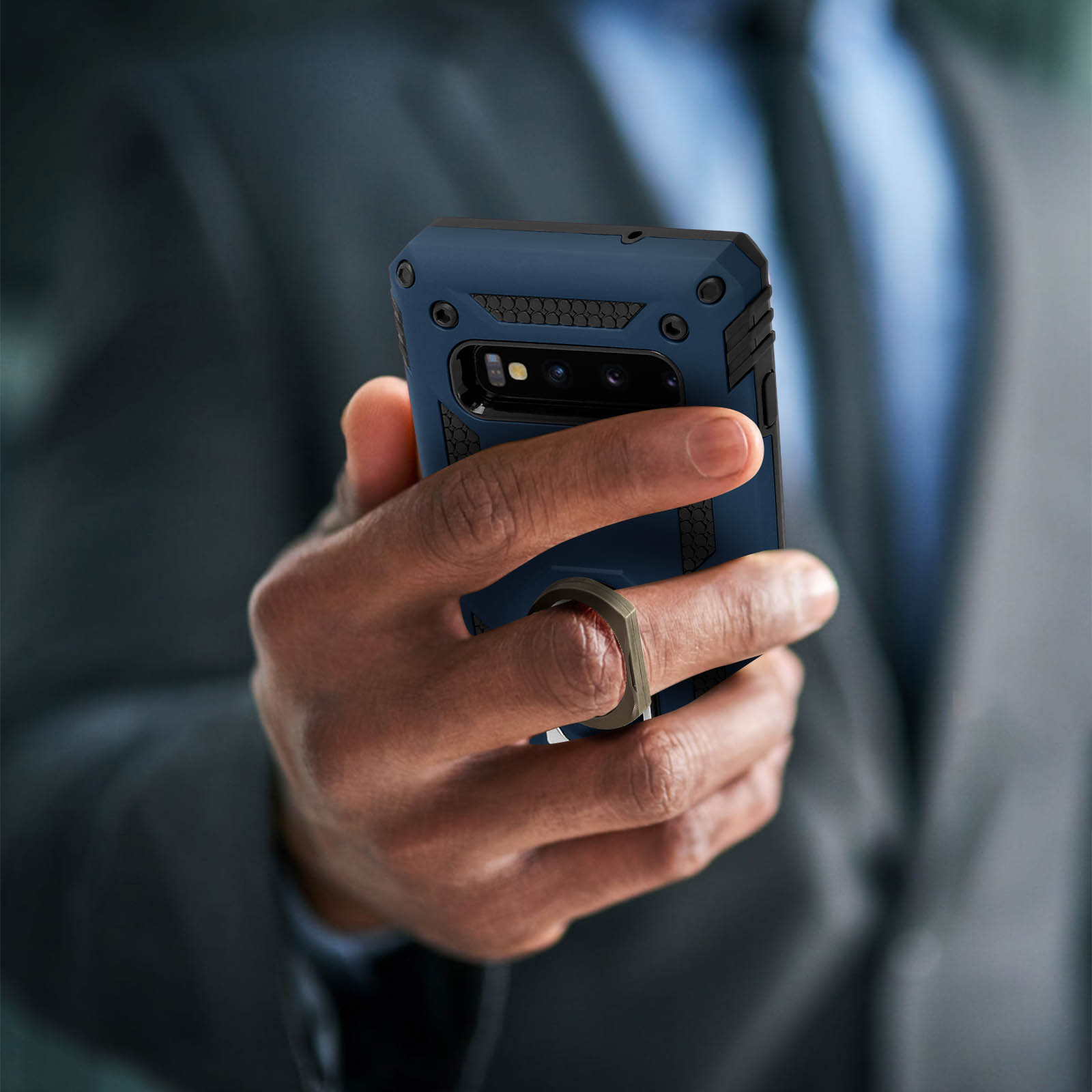 Una mano tiene un telefono in una custodia blu con un anello. Lo sfondo è un uomo in giacca e cravatta.